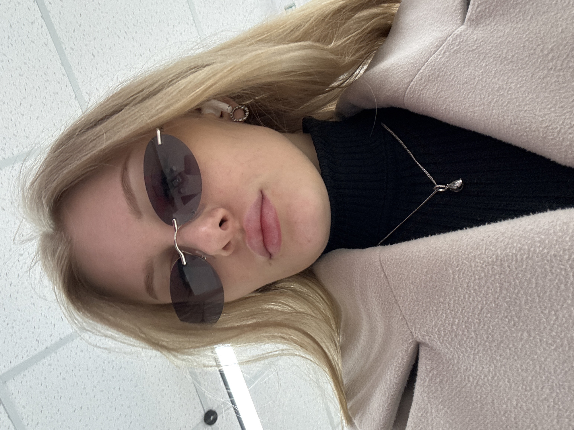 A close-up portrait of a young woman, rotated 90 degrees counter-clockwise, depicts her looking downward towards the camera. She has blonde hair and fair skin. Her eyes are obscured by rimless, dark-tinted circular sunglasses. Her lips are full and slightly parted, revealing their natural pink hue. A small, circular, possibly jeweled earring is visible in her right ear, accompanied by a white earbud.
She is dressed in a black ribbed turtleneck or sweater, over which she wears a light beige or cream-colored coat with a noticeable texture, resembling wool or fleece. A delicate silver chain necklace with a small, pear-shaped pendant rests against her black top.
The setting appears to be indoors, with a white ceiling featuring acoustic tiles or a similar patterned surface in the background. Reflections in her sunglasses and on the ceiling suggest overhead lighting fixtures, indicating a well-lit environment such as a commercial or public building. The activity captured is a self-portrait. Her expression is neutral. No text is visible within the image. The location context for this media file is Perm, Russia.