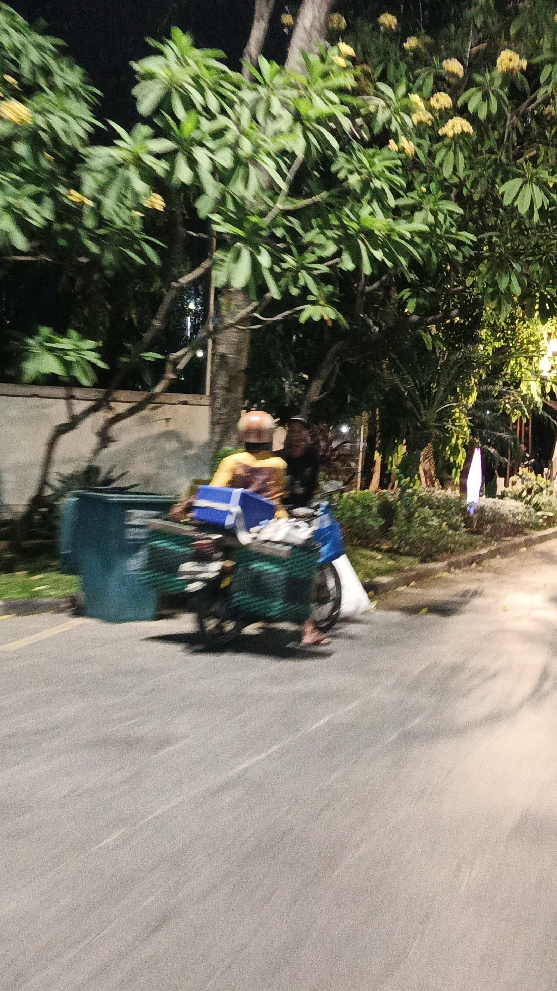 A person wearing a yellow shirt and a brown helmet is riding a motorcycle with a green basket and a blue box on the back. They are moving from left to right across a paved road at night. The rider is blurred due to motion. To the left of the motorcycle, there is a large teal trash bin. The background features trees with green leaves and some yellow flowers, a wall, and some bushes. The lighting suggests it is nighttime, with some artificial light sources illuminating the scene. The image is blurred overall, indicating movement of the camera or the subjects. The scene is likely a street in Surakarta, Indonesia, given the presence of common urban elements and vegetation typical of the region.