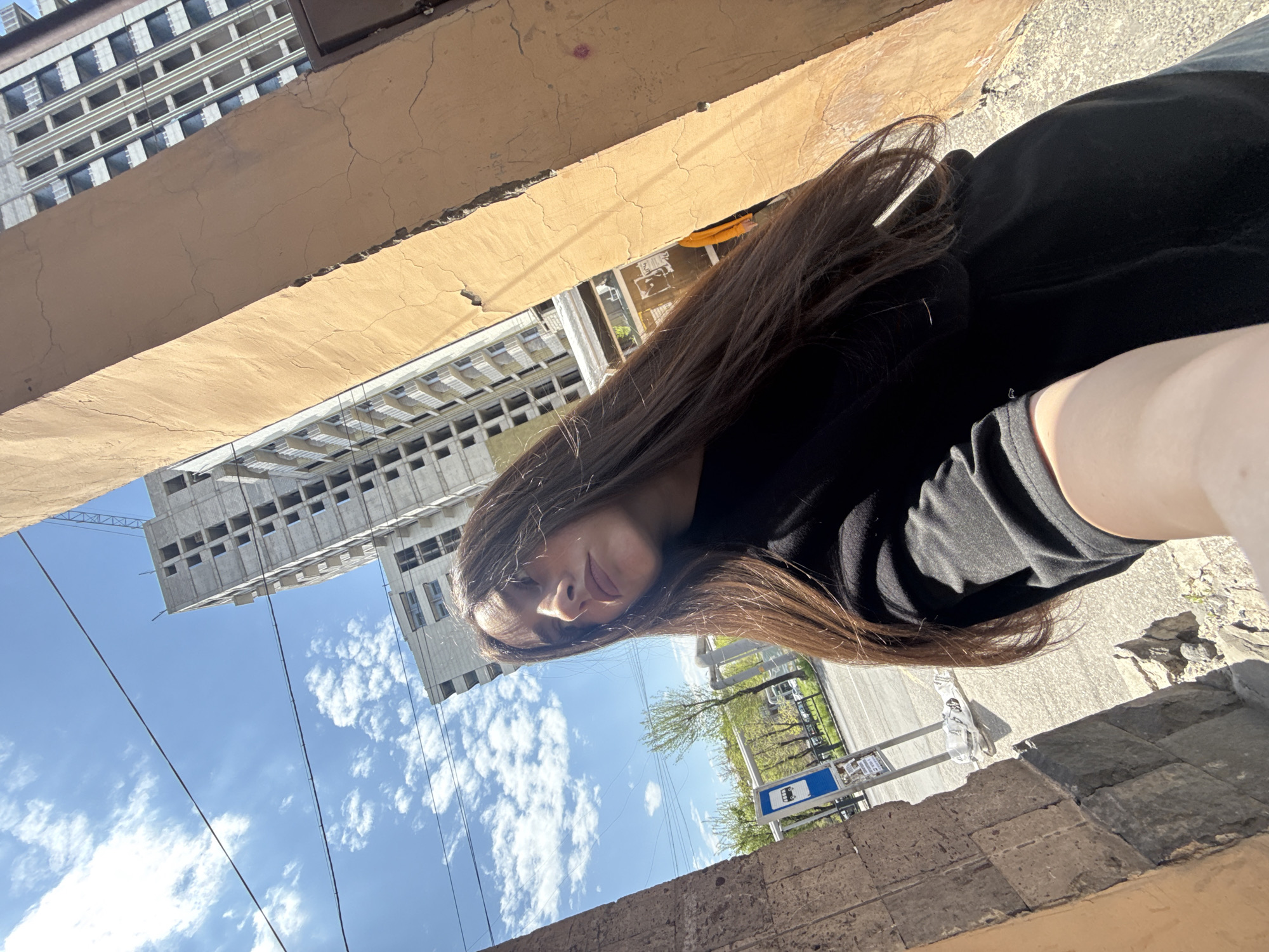 A young woman with long brown hair, wearing a black top, is taking a selfie in Yerevan, Armenia. She is standing in what appears to be an alleyway or narrow street with tall buildings on either side. One building is under construction, with exposed concrete and window frames. The sky is blue with scattered clouds. A bus stop sign is visible in the background. The surrounding architecture is a mix of old and new, with some buildings showing signs of disrepair.