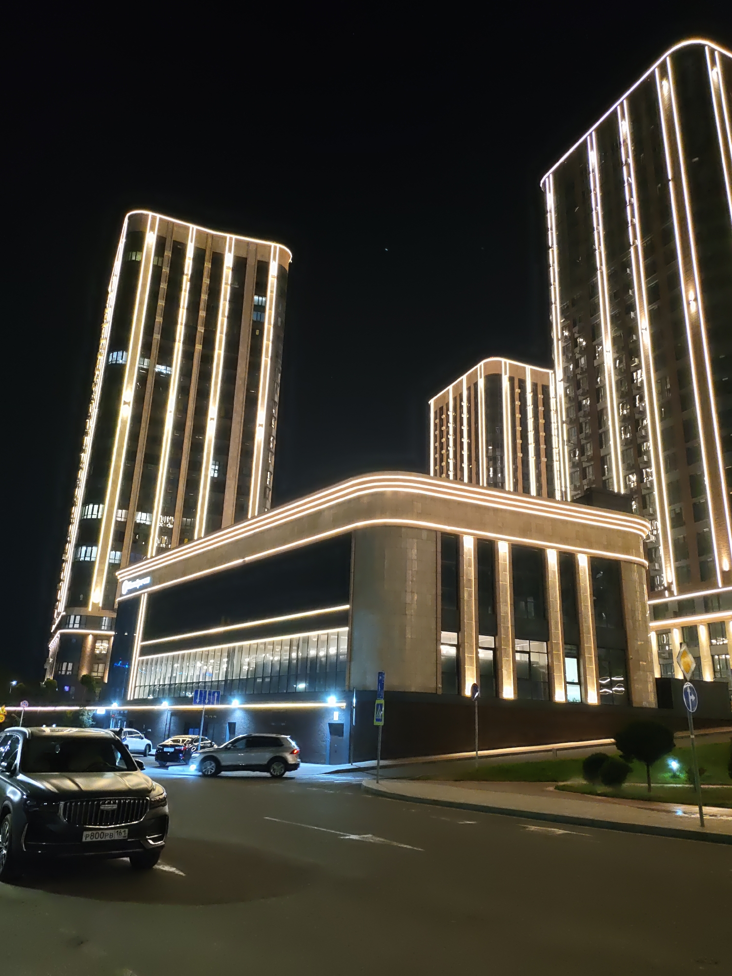 The image shows a modern urban landscape at night. The main subjects are several illuminated buildings, likely residential or mixed-use, with vertical strips of warm yellow light accentuating their facades. In the foreground, a dark-colored SUV with a license plate reading "P800PB 161" is parked on the right side of the street. Behind it, other cars are visible, including a silver SUV and a black sedan. The street appears to be paved, with sidewalks and a grassy area bordering the buildings. Several street signs and lampposts are present. The sky is dark and clear. The scene is consistent with the architectural style and streetscape of Rostov-on-Don, Russia, particularly in areas featuring contemporary developments.