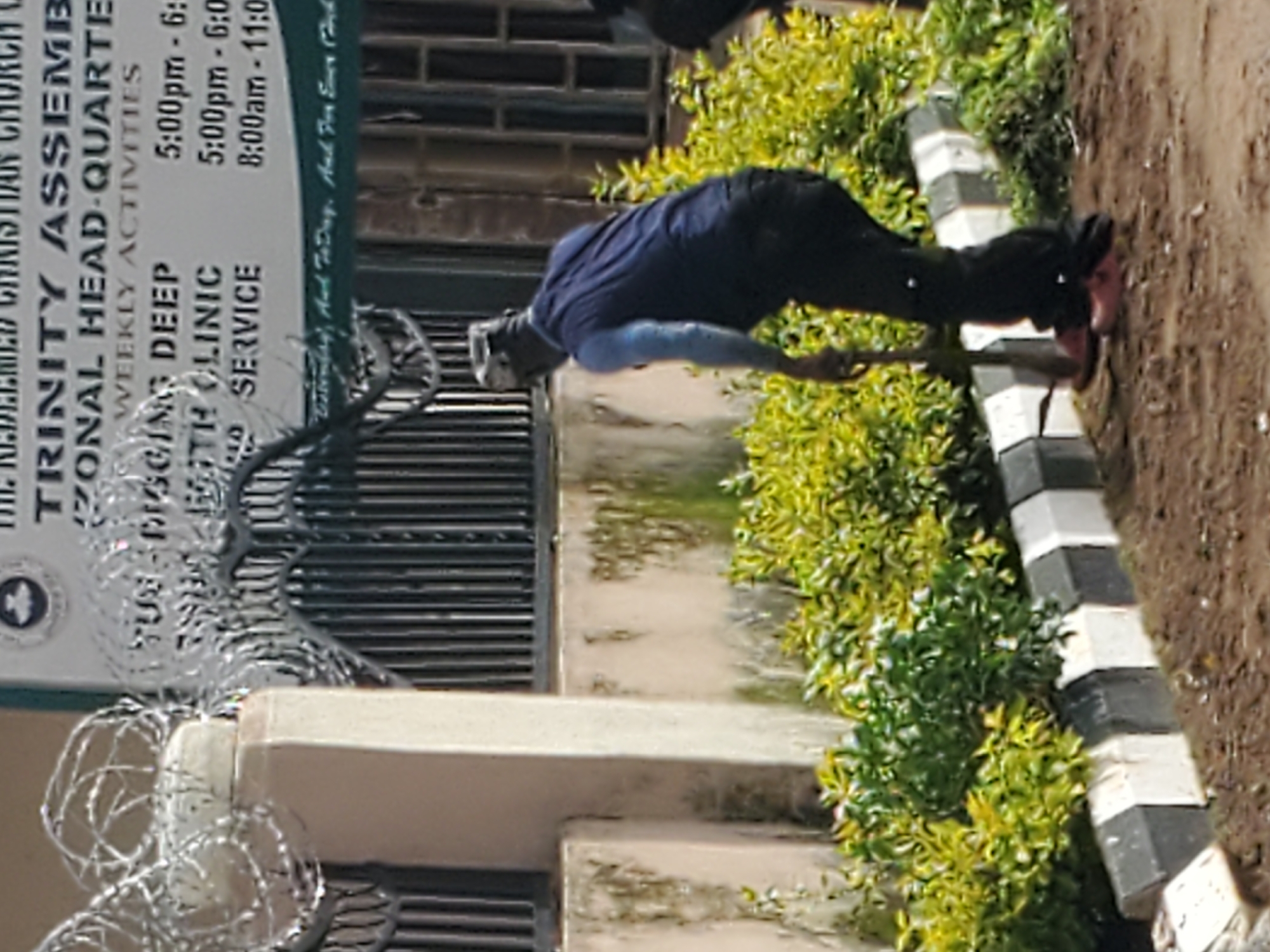 A man is tending to plants in front of a building. He is wearing a blue vest over a light blue long-sleeved shirt, black pants, and red shoes. He holds a shovel and appears to be digging or working the soil around the plants. The building behind him has a sign with text that is partially legible. The sign indicates "TRINITY ASSEMBLY ZONAL HEAD-QUARTER" and lists "WEEKLY ACTIVITIES" with times. To the left of the building, there is a concrete pillar with barbed wire wrapped around its top. The area is outdoors, with soil and a curb separating the planted area from a pathway or road. The overall scene suggests a maintenance or gardening task at the church. The image is taken during the day, with natural lighting.