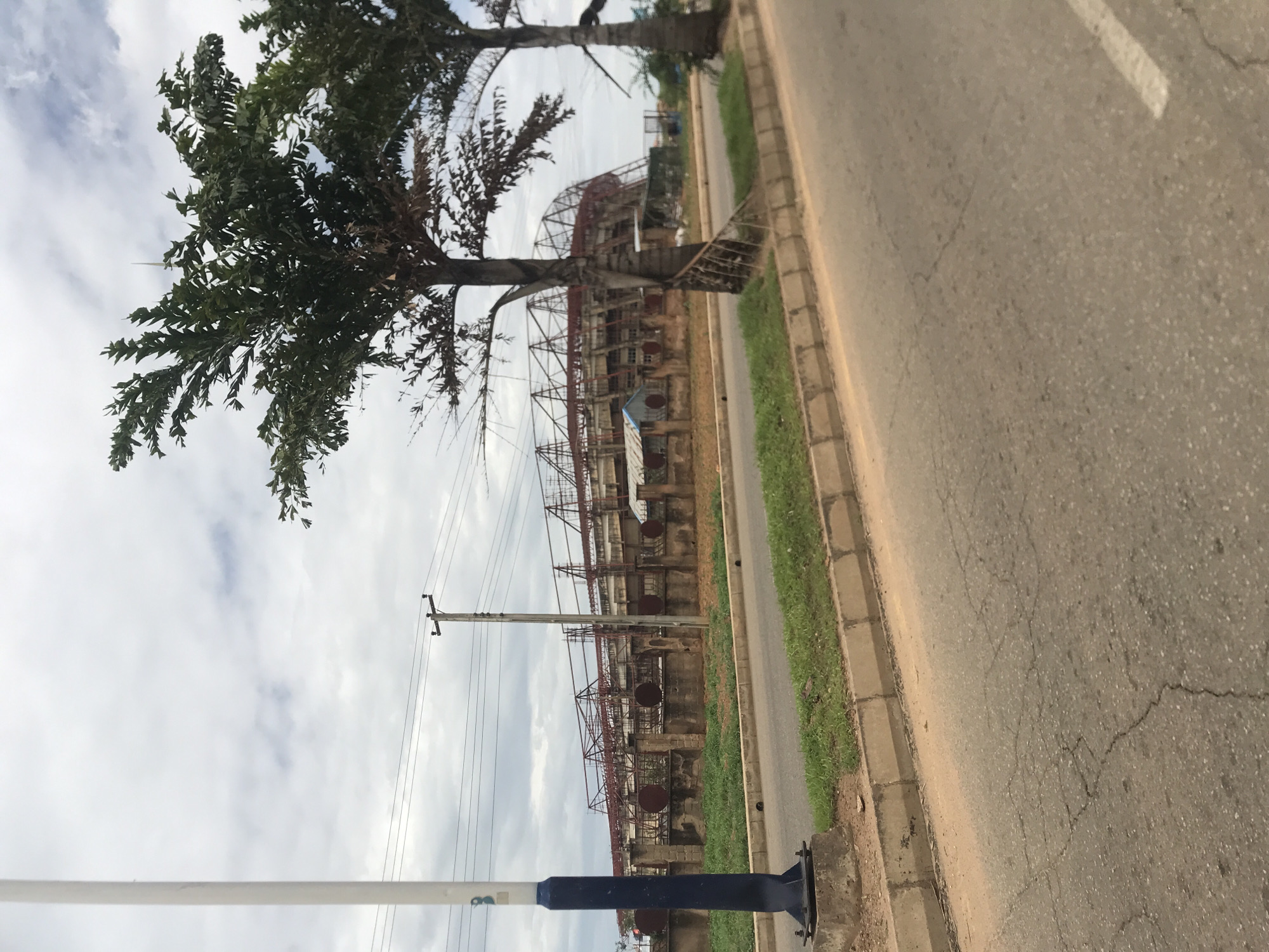 The image depicts an outdoor scene in the vicinity of the New Stadium, Nigeria, under an overcast sky. A two-lane asphalt road with visible cracks runs horizontally across the lower half of the frame. A concrete curb separates the road from a narrow, irregularly grassed median strip.
On the far left, a tall, white light pole with a blue and white painted base is anchored to a concrete footing. In the midground, a dark wooden utility pole with multiple insulators supports several power lines that stretch across the scene. Further right, two palm-like trees with textured trunks and dark green foliage stand between the road and a perimeter fence.
Beyond the median strip and trees, a concrete wall topped with an iron fence defines the boundary of a large facility. Behind this fence, several small, simple structures with corrugated iron roofs, some appearing as shacks, are visible. The dominant background feature is a large, multi-tiered concrete stadium-like structure. This building exhibits an exposed reddish-brown metal framework, possibly remnants of a roof structure or extensive scaffolding, indicating either ongoing construction, significant disrepair, or partial deconstruction. No individuals or discernible actions are present in the scene.