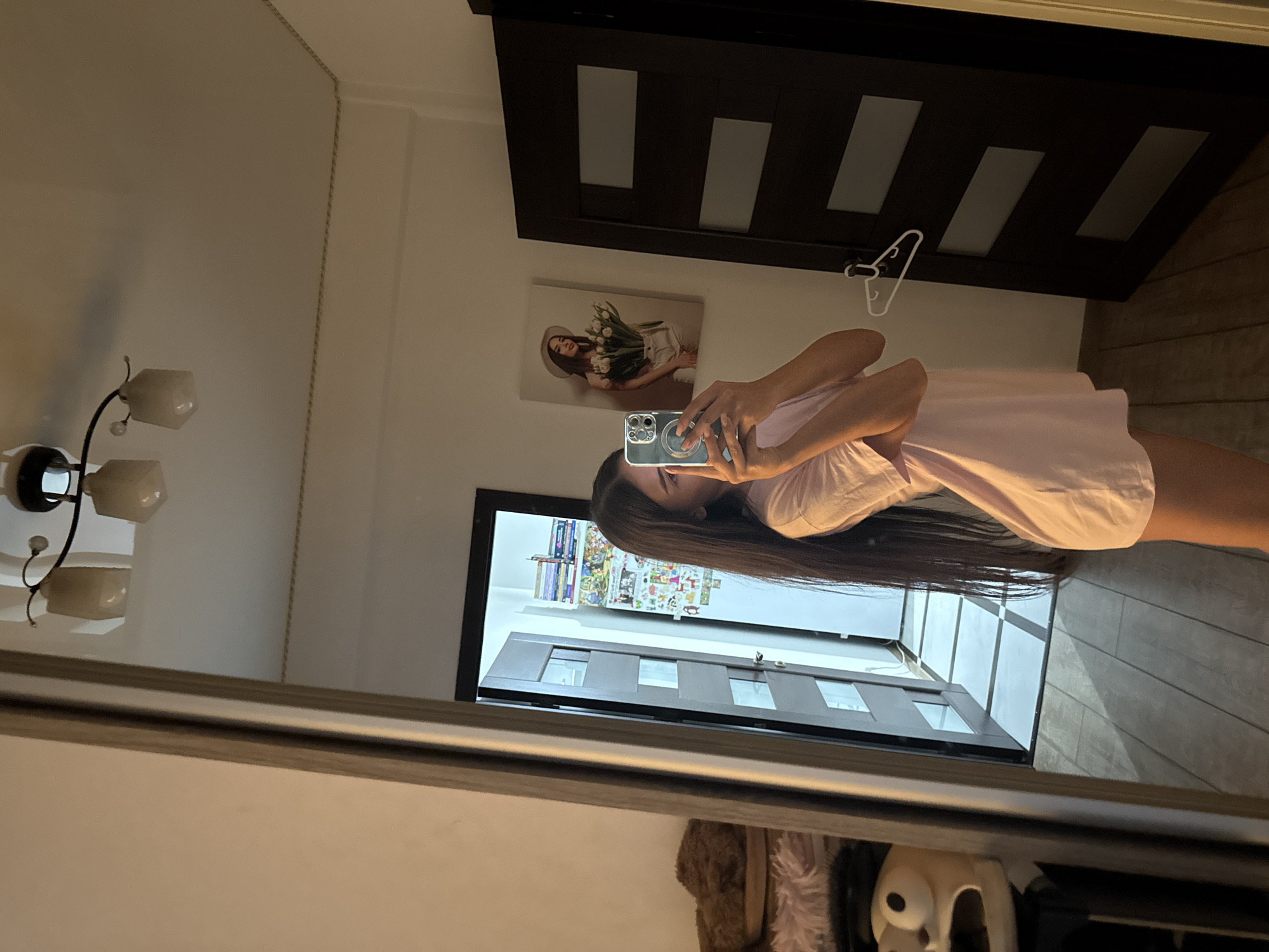 In an indoor setting, a young woman with long dark hair is standing in front of a full-length mirror and taking a selfie with her phone. She is wearing a light pink t-shirt dress and her reflection shows her holding the phone up to take the picture. The mirror reflects a room with a doorway, a painting on the wall of a woman holding flowers, and a closet door with glass panels. To the left of the mirror, part of a dresser or shelf with fuzzy brown slippers and a plush toy is visible. The flooring appears to be made of wood. The lighting suggests it is either nighttime or a dimly lit room. The location is unknown, and cannot be identified as being in Chișinău, Moldova.