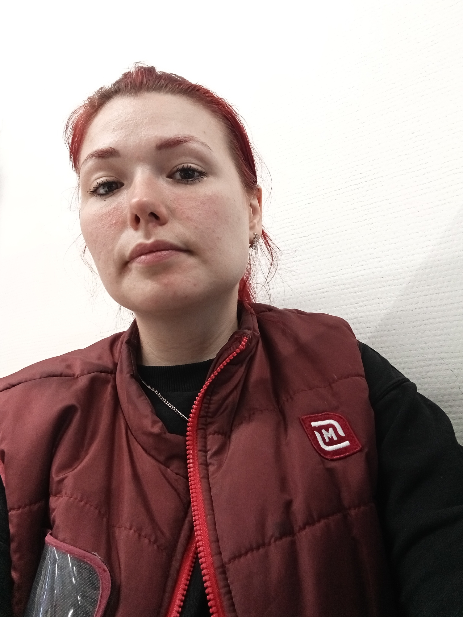 The image is a close-up, selfie-style portrait of a young woman with red hair. She is wearing a dark red quilted vest over a black top, and a silver chain necklace. A logo with the letter "M" in a square is visible on the vest. The background is a textured white wall. The lighting suggests an indoor setting. The woman's face is centrally framed, and she is looking directly at the camera. Her expression is neutral. The image does not provide any specific clues to its location in the city of Chaikovsky, Russia, or any specific actions or interactions.