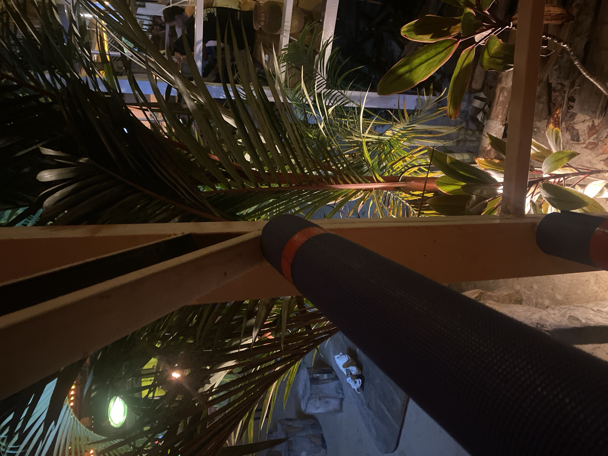 The image shows a nighttime scene in Baan Hin Kong, Thailand. The viewpoint is from a low angle, looking upwards through a balcony or raised platform structure made of light-colored metal framing. A dark-colored yoga mat with an orange band is rolled up and positioned along the railing. Lush vegetation, including palm trees and other plants with broad, colorful leaves, surrounds the structure. Behind the vegetation, there is a building with lighting.