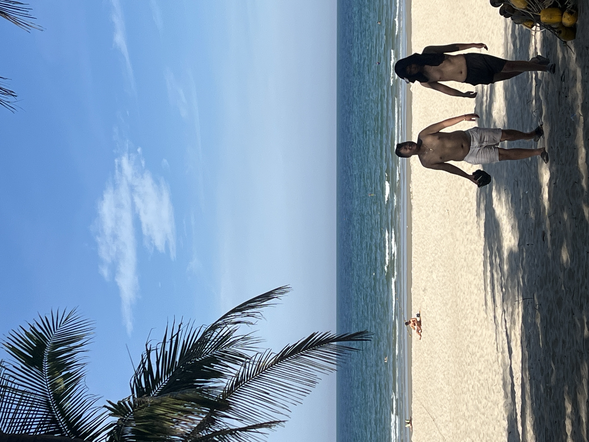 The image depicts a sunny day at a beach in Da Nang, Vietnam. In the foreground, two shirtless men are walking on the sandy shore. The man on the left wears light-colored shorts and holds a dark hat in his hand. The man on the right wears dark shorts and has a black cloth draped over his head and shoulders. In the middle ground, a person is sunbathing on the sand near the water's edge. The ocean stretches across the background, with gentle waves rolling onto the beach under a clear blue sky with scattered white clouds. Palm fronds from a tree hang over the left side of the frame, casting shadows on the sand. In the far distance, several figures can be seen in the water. To the right of the frame, a cluster of yellow buoys is visible on the sand.