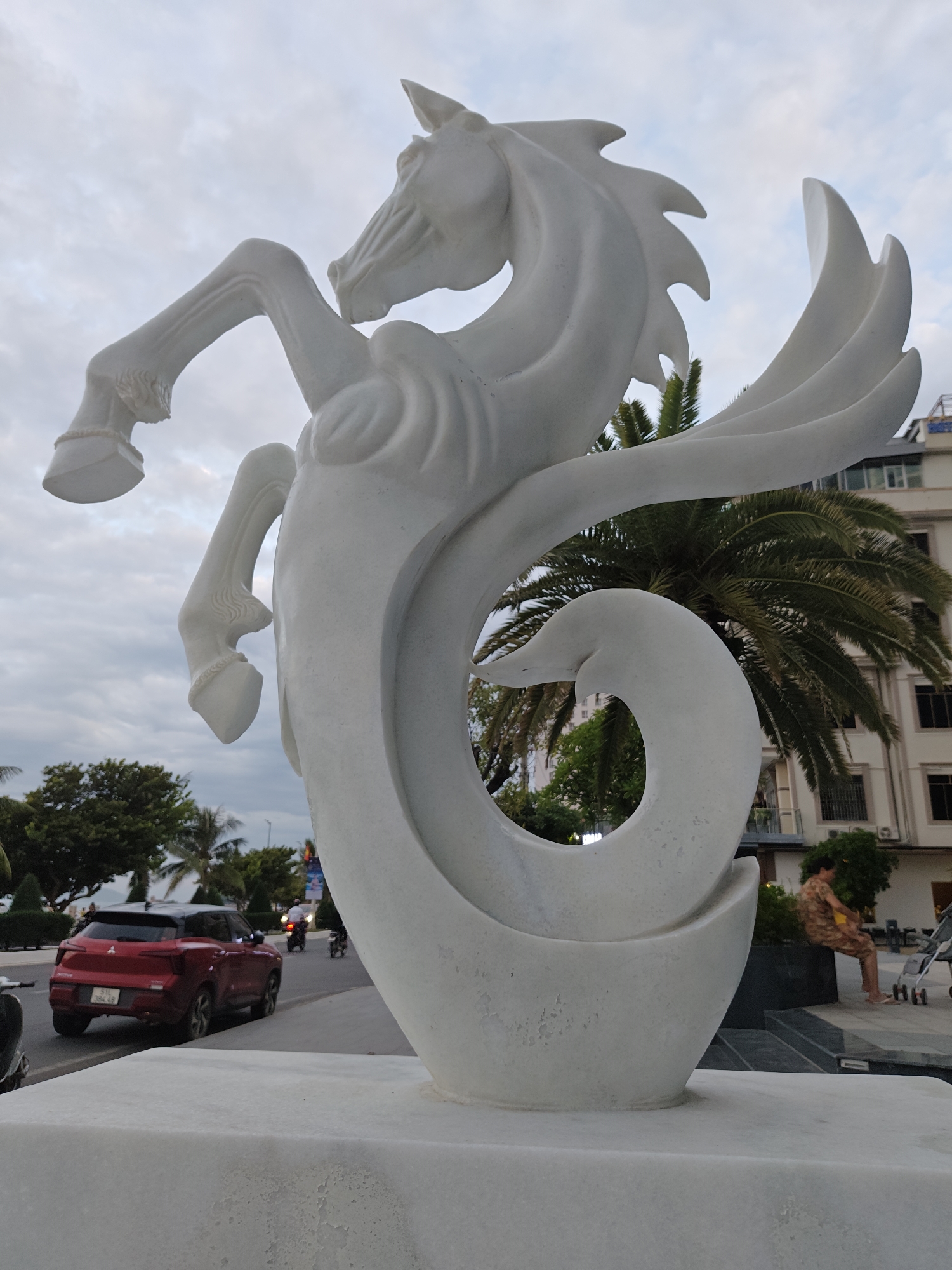 A white, abstract sculpture of a winged horse is the primary subject, positioned in the foreground against an overcast sky. The sculpture is made of smooth, polished stone and features dynamic lines, with one foreleg raised and wings spread behind its head. The lower part of the sculpture curves into a circular shape that resembles a swan's neck. Behind the sculpture, a palm tree is visible, along with a multi-story building and a street with moderate traffic. A red SUV is in the middle ground, driving on the right side of the road. Further in the background, a person is sitting on a bench, and a stroller is nearby. The scene appears to be a public outdoor space, likely a waterfront promenade or park, consistent with locations in Nha Trang, Vietnam, given the architectural style and tropical vegetation.