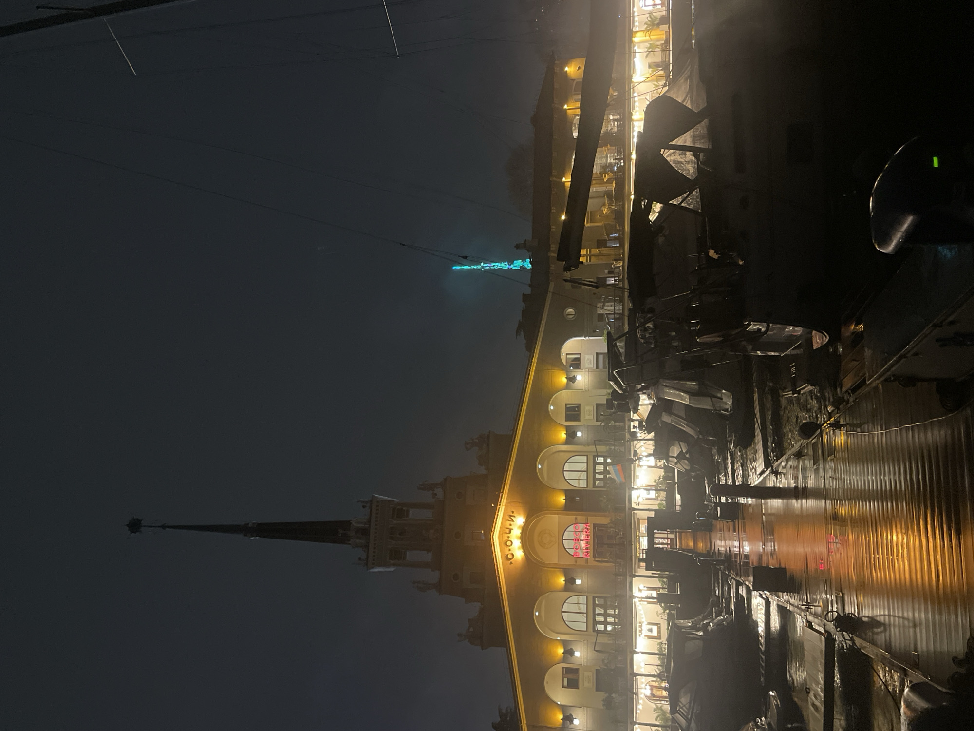 This is a night shot of the Sochi Marine Station, located in Sochi, Russia. The building is illuminated with warm yellow lights, contrasting with the dark sky. The image is taken from a low angle, possibly near the harbor, with wet surfaces reflecting the light. The Sochi Marine Station tower is visible to the left of the frame. Some masts and rigging from boats are visible on the right side.