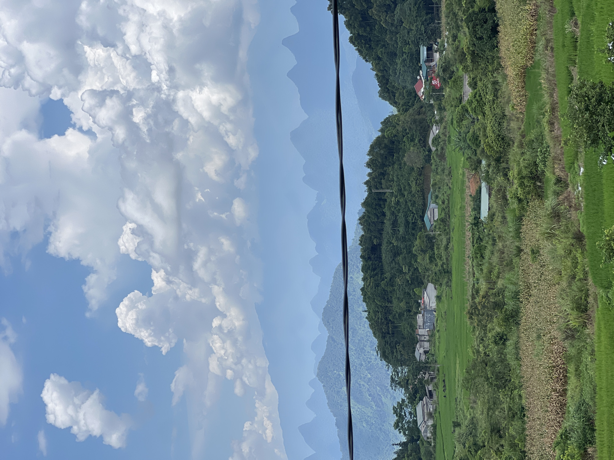 The image depicts a rural landscape in Quản Bạ Commune, Vietnam, characterized by rolling green hills, lush vegetation, and a cluster of houses nestled amidst the foliage. In the foreground, vibrant green rice paddies dominate, interspersed with dry corn stalks and other types of foliage. A small waterway is visible. Further back, a small village with several houses, some with red roofs, is scattered across the lower slopes of the hills. Trees densely cover the hillsides, with a prominent line of trees forming a ridge. In the mid-ground, more houses are visible, some with light-colored walls and tile roofs, situated on a higher elevation. The background is dominated by a series of majestic, mist-shrouded mountains, their peaks fading into the hazy blue sky. The sky itself is a bright blue, adorned with scattered, fluffy white clouds. A dark, horizontal power line stretches across the mid-section of the image, obscuring a portion of the mountainous scenery. The overall scene conveys a sense of peaceful, rural life within a dramatic natural setting. No people or specific actions are discernible in the image.