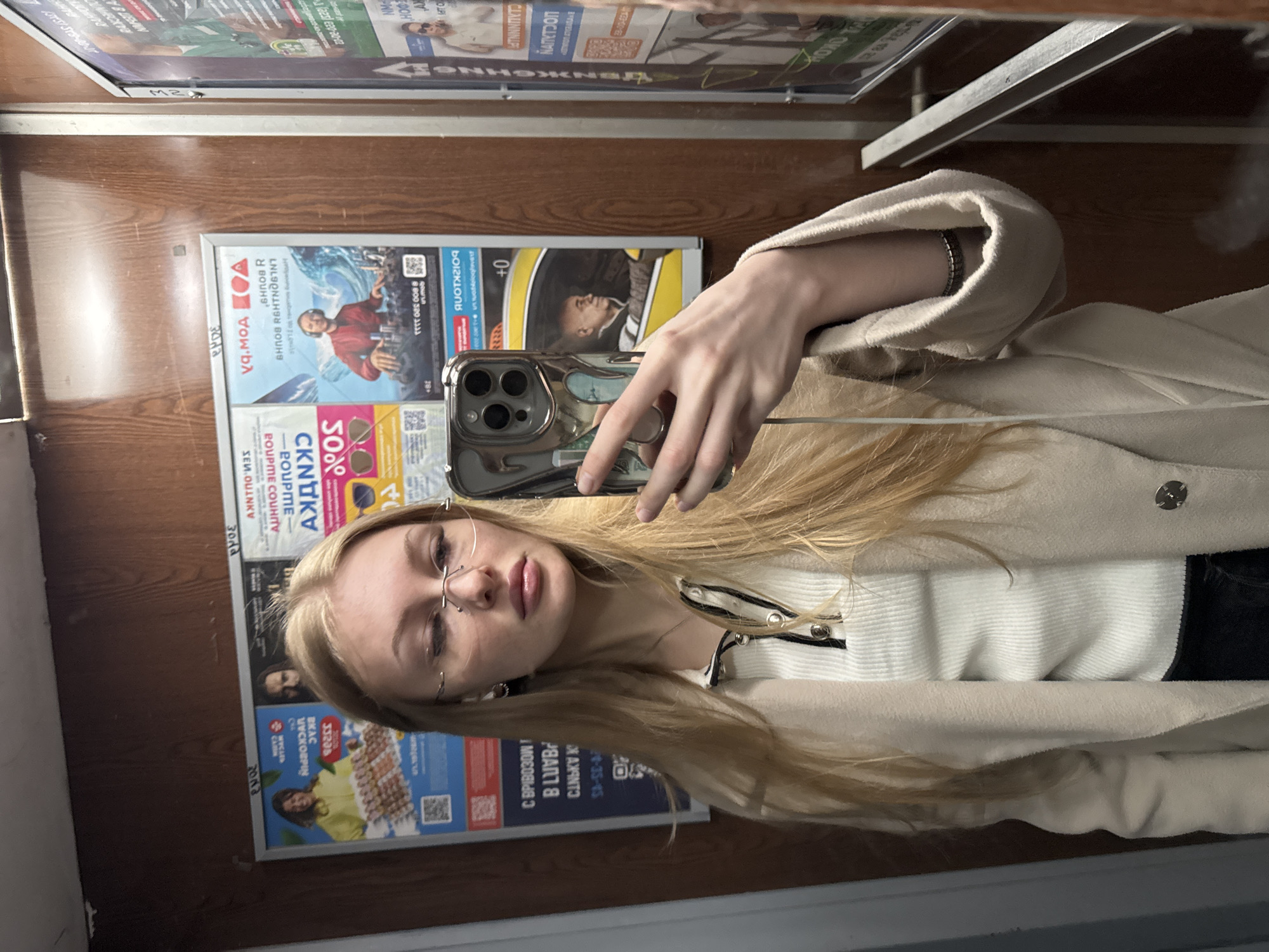 A young woman with blonde hair and glasses is taking a selfie with a smartphone. She is wearing a white top and a beige jacket. Behind her is a wall with several advertisements posted on it. The advertisements are in Russian, and some have QR codes. The location is likely a public transportation station in Perm, Russia.
