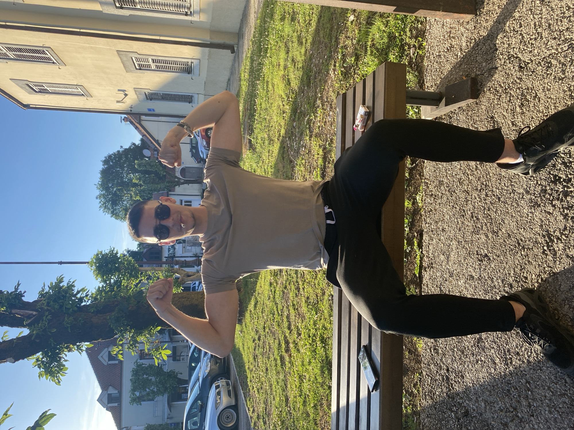 A young man is sitting on a wooden bench in a park in Cetinje, Montenegro. He is wearing a beige t-shirt, black sweatpants, and black sneakers. He is flexing his biceps, showing off his muscles. He is also wearing sunglasses and has a cigarette in his mouth. On the bench next to him, there is a smartphone and a pack of cigarettes. The park is green and grassy, with trees and buildings in the background. The sun is shining, and the sky is blue.
