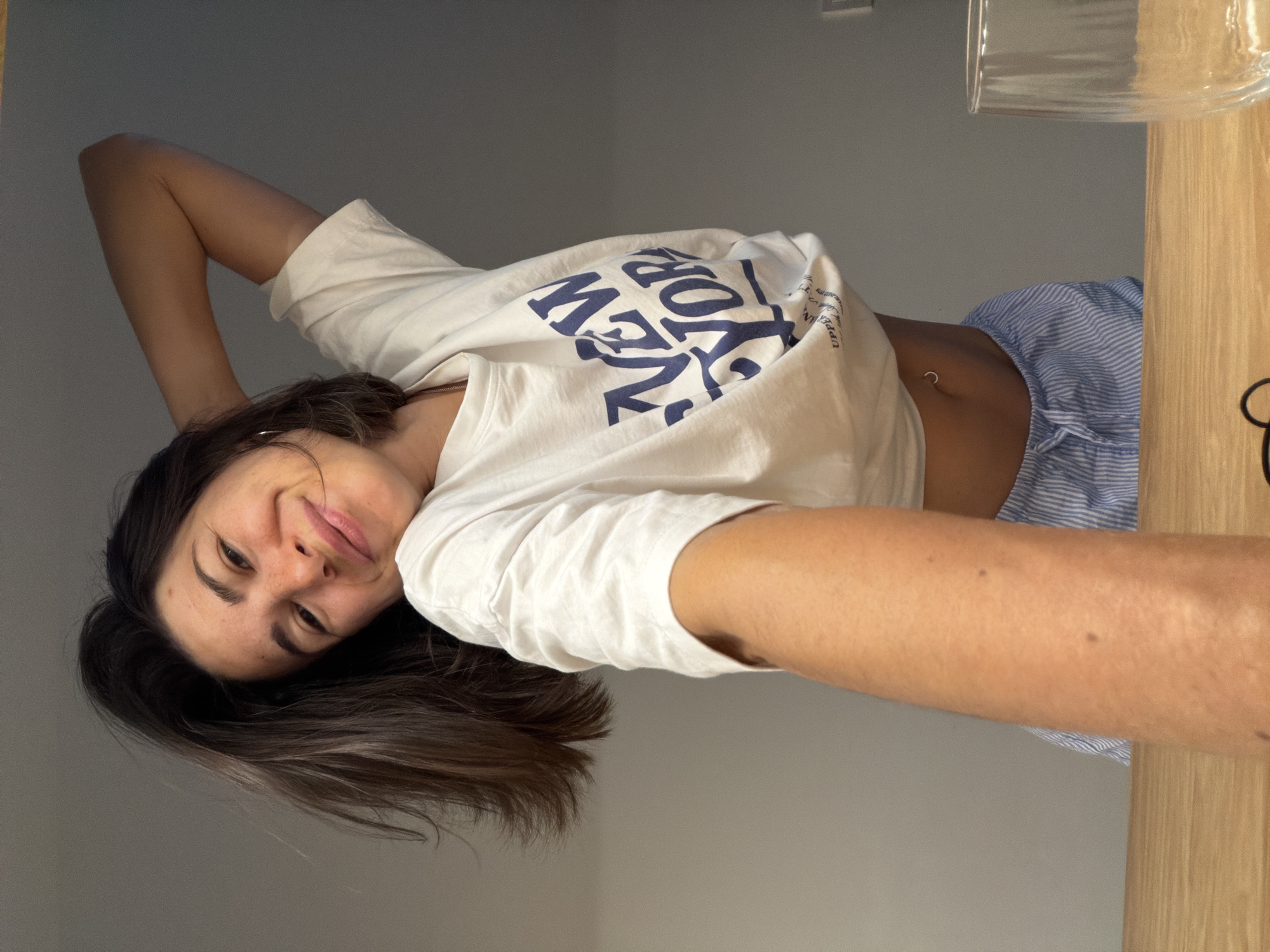 A young woman is seen in a selfie-style photo. She is wearing a white t-shirt with "New York" printed on it in blue letters, as well as blue and white striped bottoms. She is smiling and looking at the camera. The location is not identifiable as Budva, Montenegro. The background includes a gray wall and a wooden surface. A glass object is partially visible in the upper right corner.