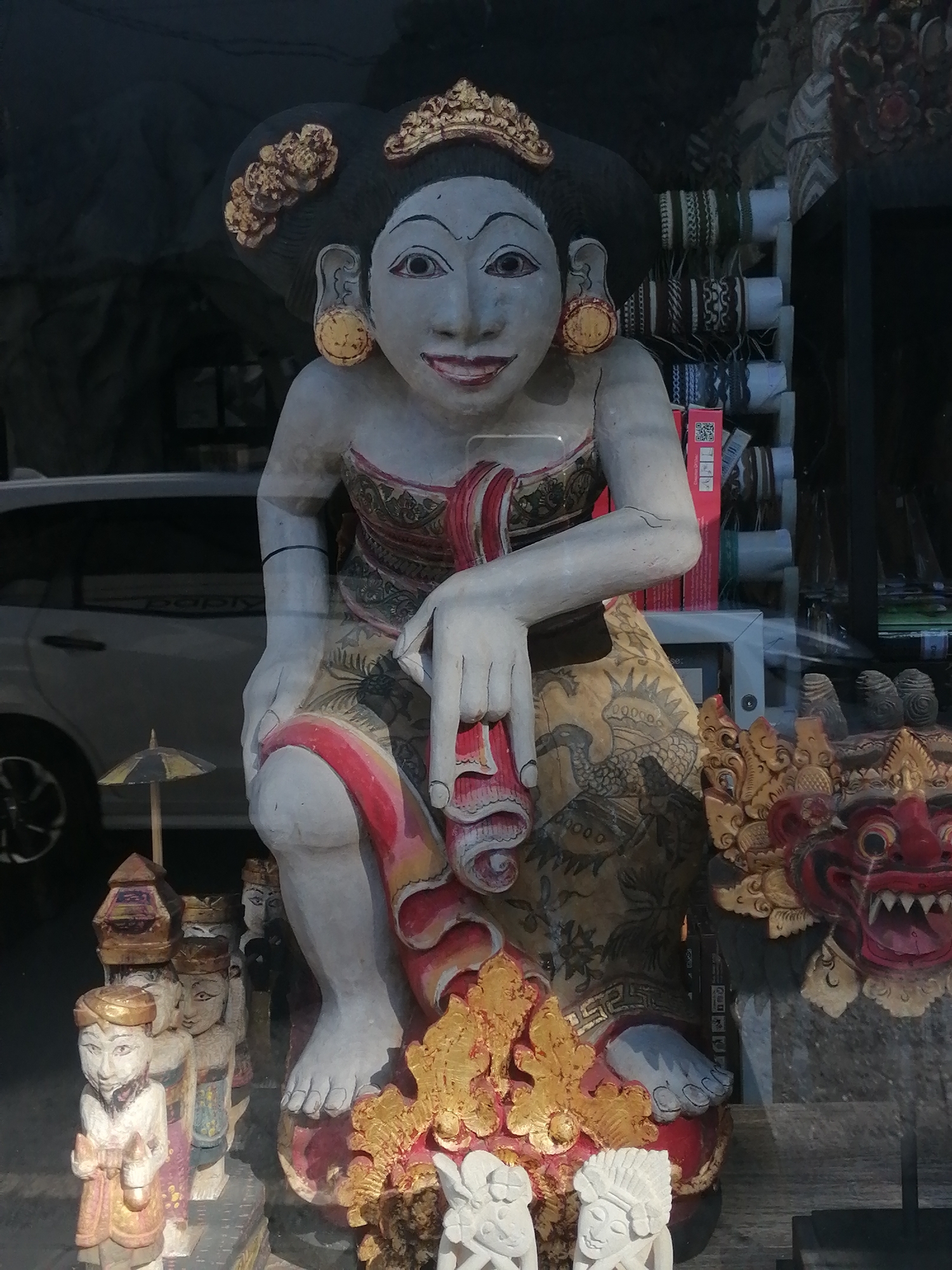 The image shows a display of Balinese art and craft items, likely in a shop or market stall in Ubud District, Indonesia. A large, full-body statue of a woman, carved and painted, is prominently featured in the foreground. She has dark hair styled with golden ornaments and wears traditional clothing with intricate patterns. Her expression is serene, and she holds a rolled-up fabric in her hand.
To the left of the main statue, there are several smaller wooden figures arranged in a row, each appearing to be a male character with different headdresses and clothing. One of these figures is holding a miniature umbrella.
To the right of the main statue, a colorful, carved mask with a fierce expression and red and gold accents is visible. Behind these objects, a white car can be seen through a window or glass, and in the background, shelves are stocked with various items, possibly textiles or other handicrafts. The overall setting suggests a retail environment selling traditional Balinese art.