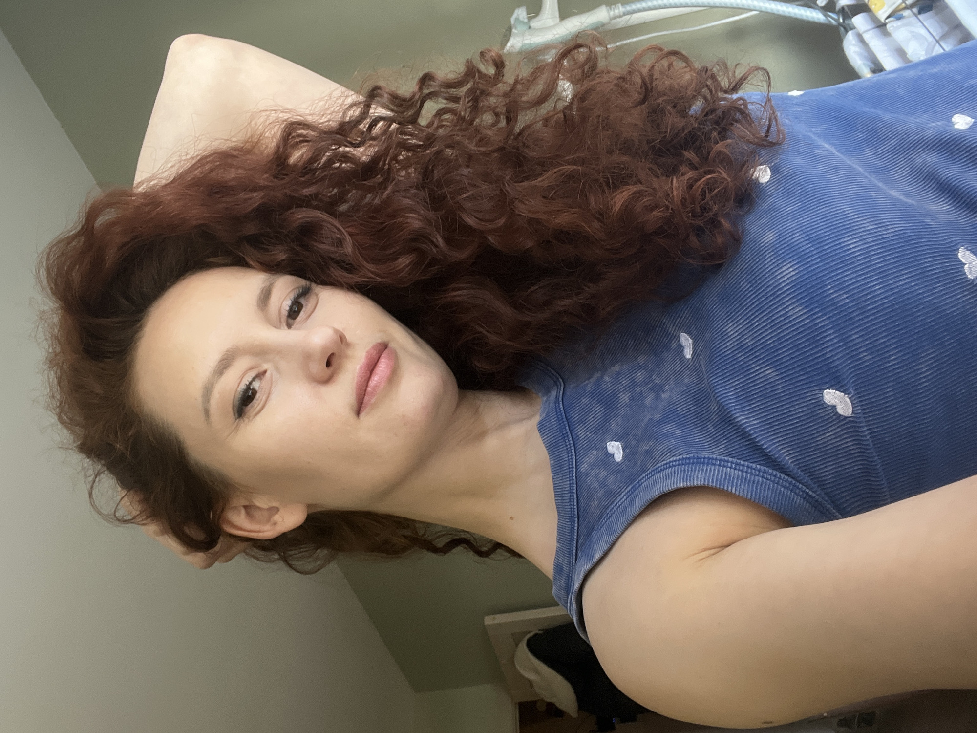 A woman with reddish-brown curly hair is lying down. She is wearing a blue corduroy shirt with small white heart patterns. Her right arm is raised behind her head. The background consists of a light green wall and some indistinct plumbing fixtures.