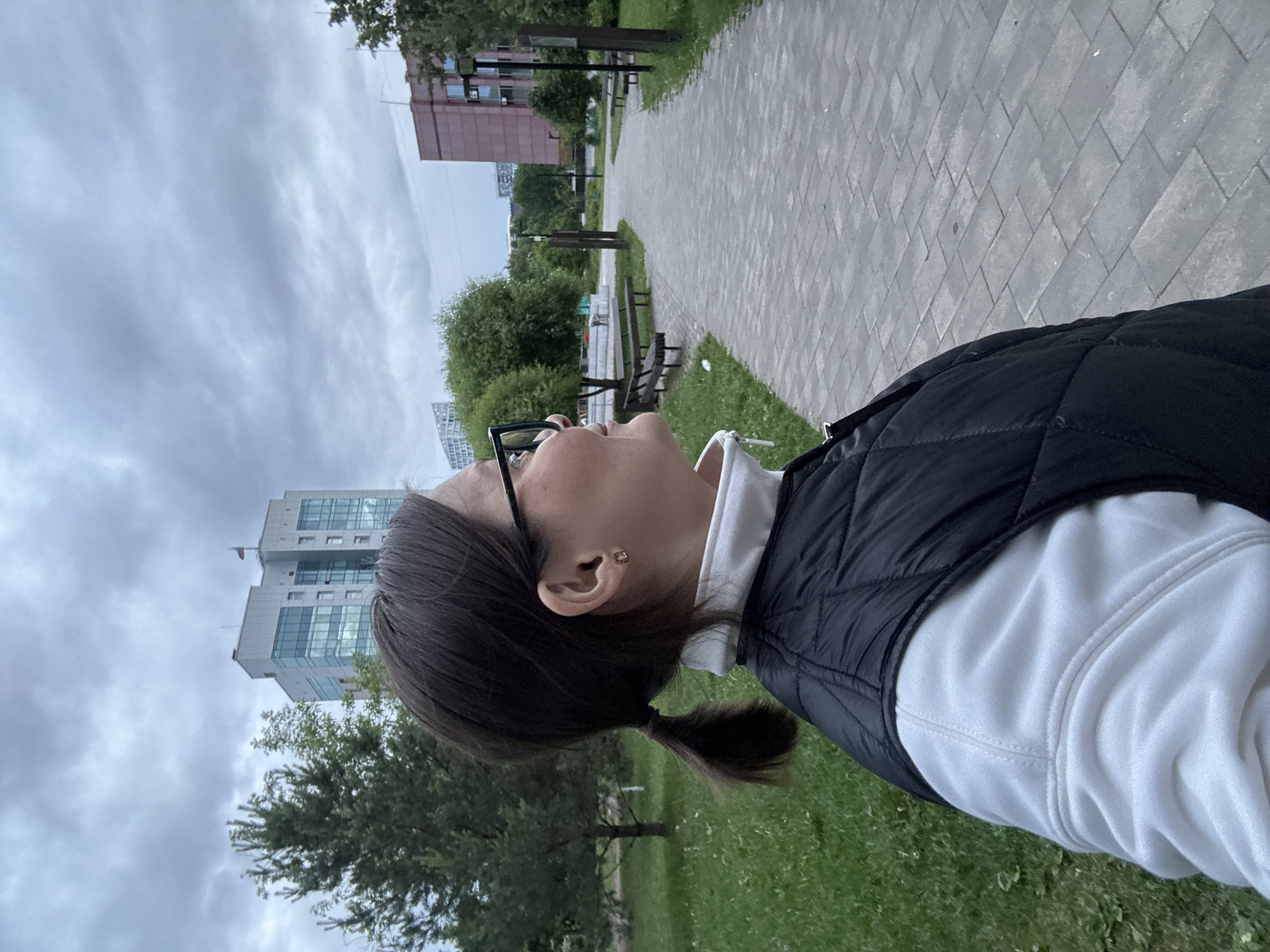 An outdoor, eye-level, profile shot on a cloudy day shows a woman looking toward the right. She has dark hair styled in a low ponytail, wears black-framed glasses, a white high-collared garment, and a black quilted vest. A small, gold-colored earring is visible in her right ear. The woman occupies the left foreground, her gaze directed along a grey paved path that stretches into the mid-ground. The path is bordered by green grass and scattered trees, with multiple dark wooden benches and streetlights positioned along its length. In the left background, a prominent, modern, multi-story building with a light-colored facade and extensive glass panels is visible, with a Russian Federation flag flying from its roof. This building is identified as the Government House of the Udmurt Republic in Izhevsk, Russia. To the far right in the background, a portion of a reddish-brown building is also visible. The sky is completely overcast.