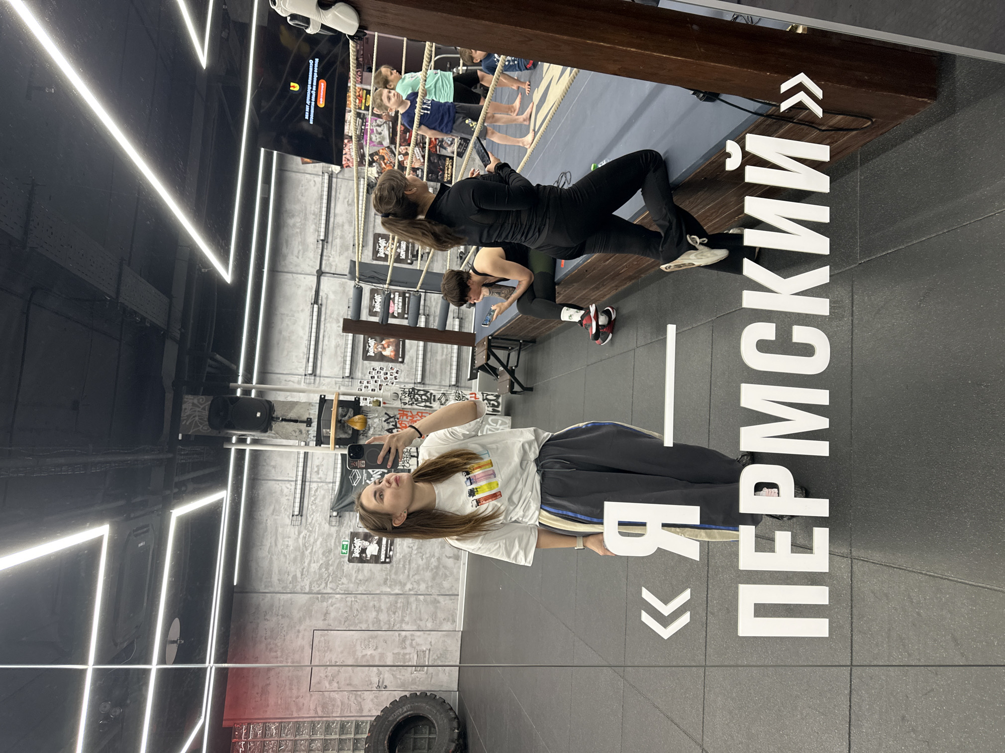 A young woman takes a selfie in front of a reflective surface with "Я - ПЕРМСКИЙ" in large Cyrillic letters (meaning "I am Permian") on the reflective surface. The location appears to be a gymnasium with workout equipment. The woman is wearing a white t-shirt with colorful designs and dark-colored sweatpants. A second reflection shows two women near a boxing ring, one holding a phone. A monitor above the ring shows a view of the ring with children. The reflections are of the gym interior. A tire is seen at the bottom of the image. The image is taken inside a location within the city of Perm, Russia.