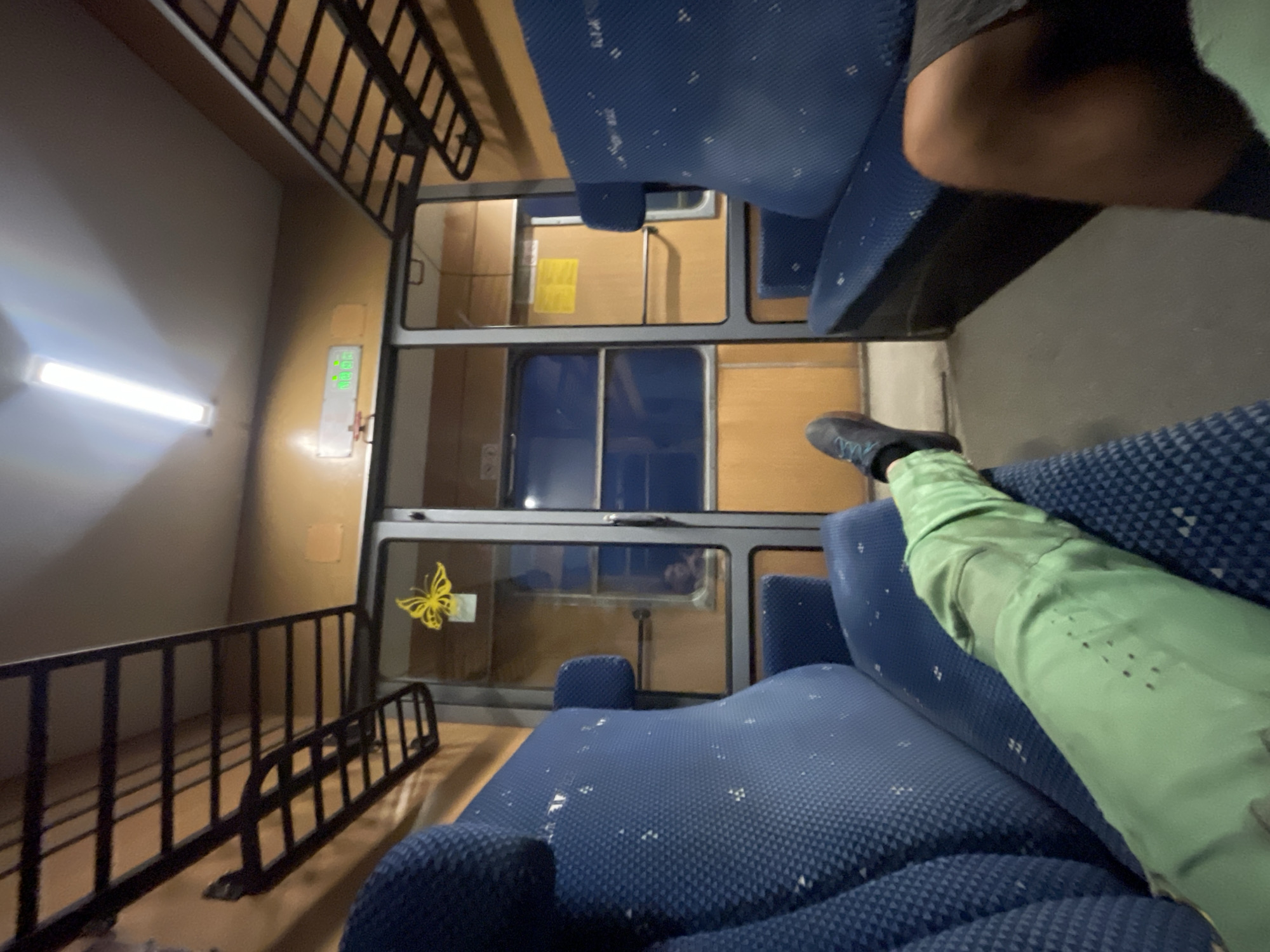 The image captures the interior of a train carriage. Blue upholstered seats line both sides of the aisle, with overhead luggage racks made of black metal. A yellow butterfly sticker is affixed to the glass door separating compartments. A bright fluorescent light illuminates the ceiling. A person's legs, clad in green pants, are visible in the foreground, resting on a seat. Another person's leg, wearing dark shorts, is seen on the right. The train carriage appears to be in motion, as suggested by the slightly blurred details. The scene is generic and does not provide any specific clues to its location within Borovo, Bulgaria.