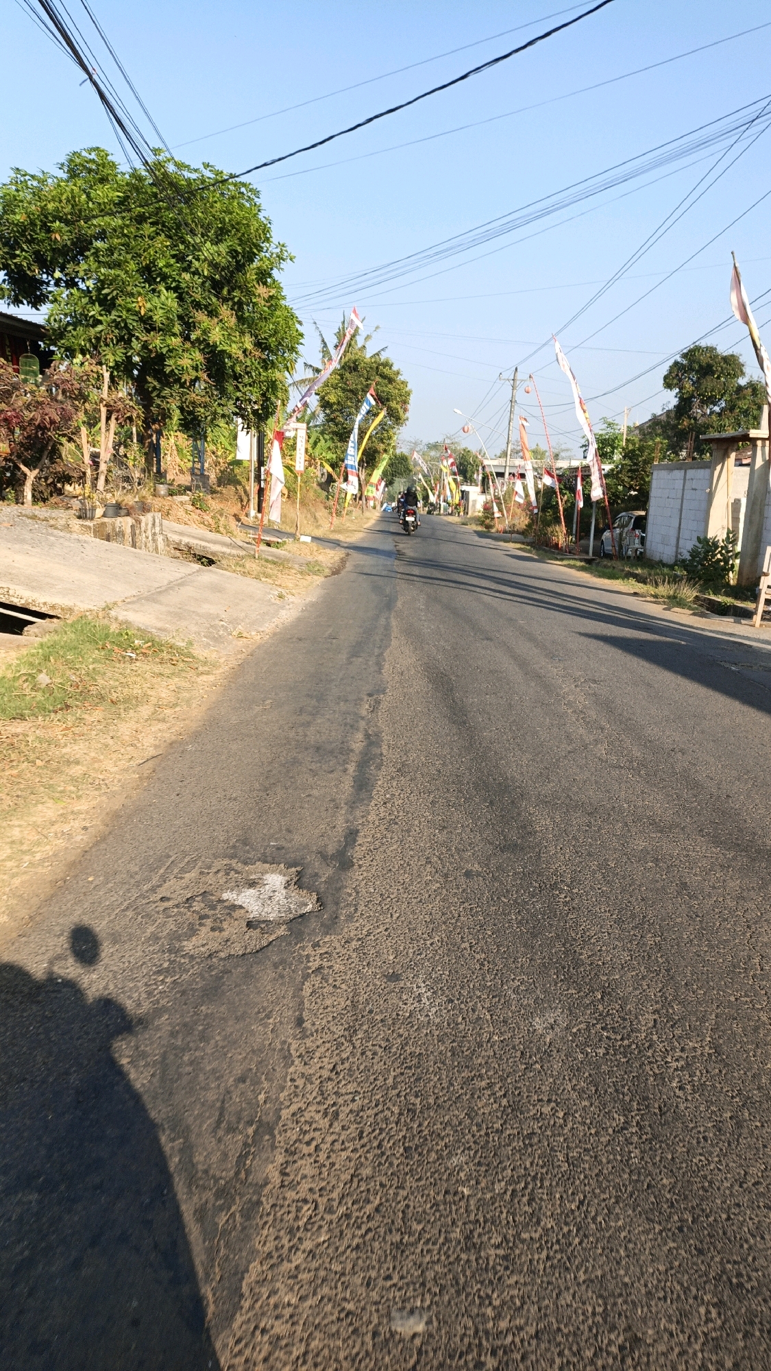 A street scene in Kuwasen, Indonesia shows a paved road with a motorcycle rider casting a shadow on the left side of the frame. The road is lined with trees, houses, and numerous Indonesian flags on poles. The sky is clear and blue, indicating it is daytime.