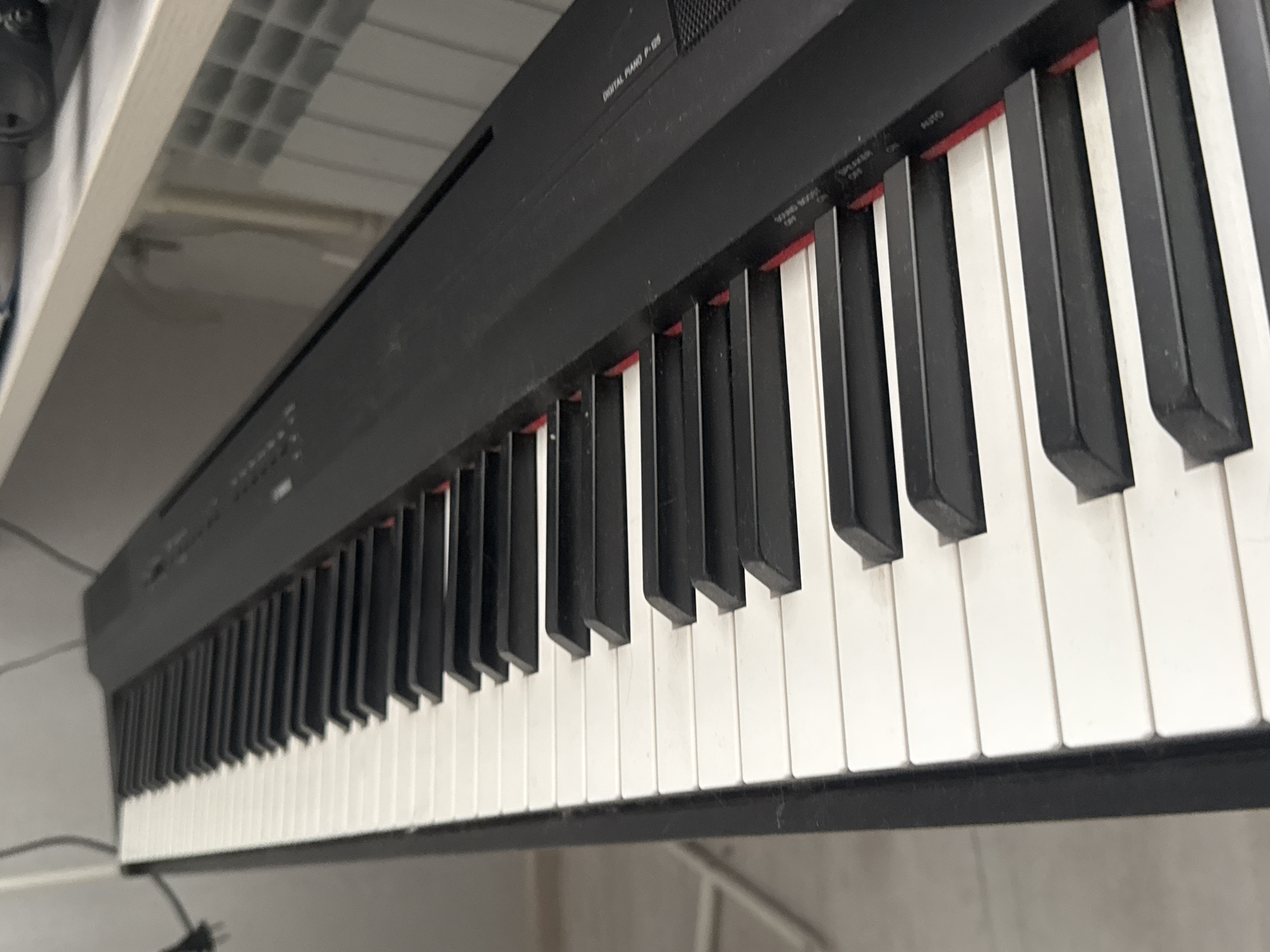 The image shows a close-up, angled view of a black digital piano with white keys. The piano is oriented diagonally from the lower left to the upper right of the frame. Some of the keys are slightly dusty. Text is visible on the right side of the piano's body, including "DIGITAL PIANO P-125" and other smaller labels that are not entirely legible. To the right of the piano, a light-colored surface is visible, possibly a shelf or desk, with a portion of a white radiator with square openings behind it. The background to the left is out of focus and appears to be a wall and some equipment, including a black cable. There are no people or animals present in the image. The scene appears to be an indoor setting. The image does not provide any specific visual cues to indicate its location in Nizhny Novgorod, Russia.