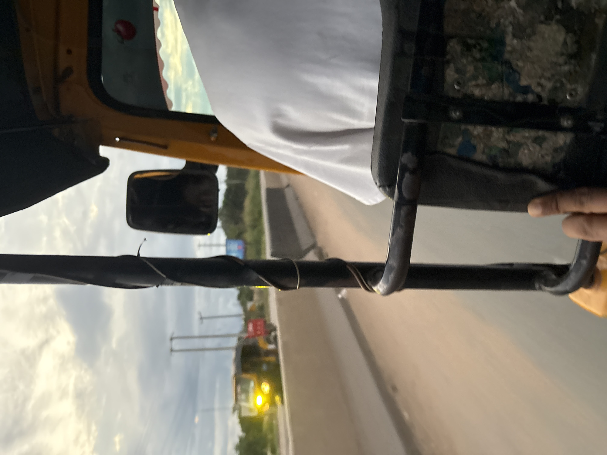 The image depicts a view from inside a yellow auto-rickshaw (Keke Napep) traveling on a road in Onitsha, Nigeria. The foreground features a person's hand resting on a black metal bar, part of the auto-rickshaw's structure. The person is wearing a white t-shirt. The auto-rickshaw is yellow and appears to be in motion, as suggested by the blurred road and background. In the mid-ground, another auto-rickshaw with its headlights on is visible on the same road, alongside a red vehicle. The road is bordered by a concrete barrier on the left, with vegetation and utility poles visible in the distance under a cloudy sky. The overall scene captures a typical street view from within a public transport vehicle in a Nigerian city.
