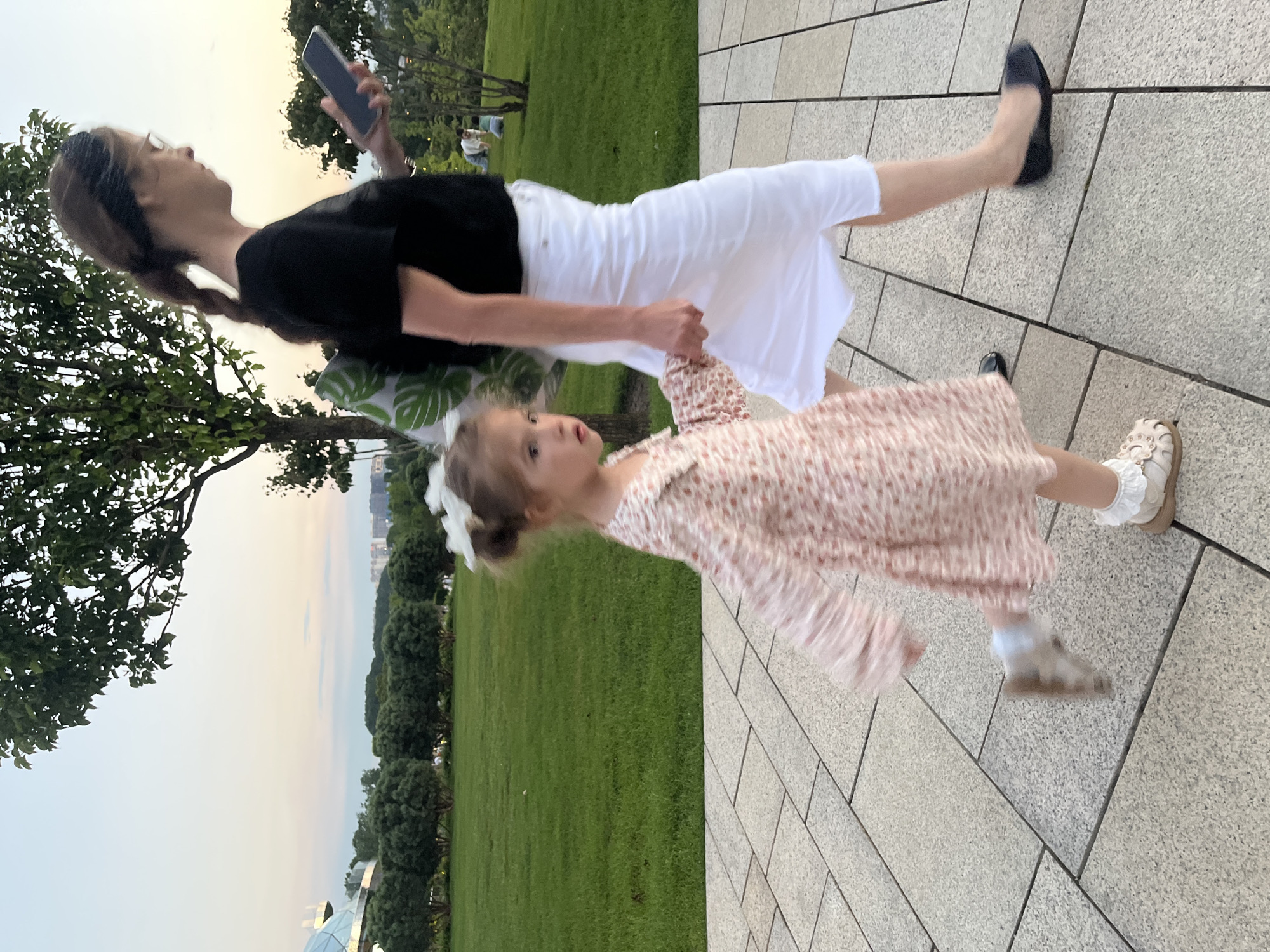 In Krasnodar, Russia, a woman and a young girl are walking on a paved pathway in a park. The woman, with her hair in a braid and wearing glasses, is dressed in a black t-shirt and a white skirt, and is looking at her phone. She has a backpack with a leaf pattern slung over her shoulder. The girl, adorned with a white bow in her hair, wears a floral print dress and light-colored sandals. She is holding the woman's hand and appears to be walking ahead of her. The background features a lush green lawn, trees, and in the distance, buildings and a building with a domed roof, suggesting an urban park setting. The sky exhibits a soft pastel hue, indicating it is likely dusk or dawn.