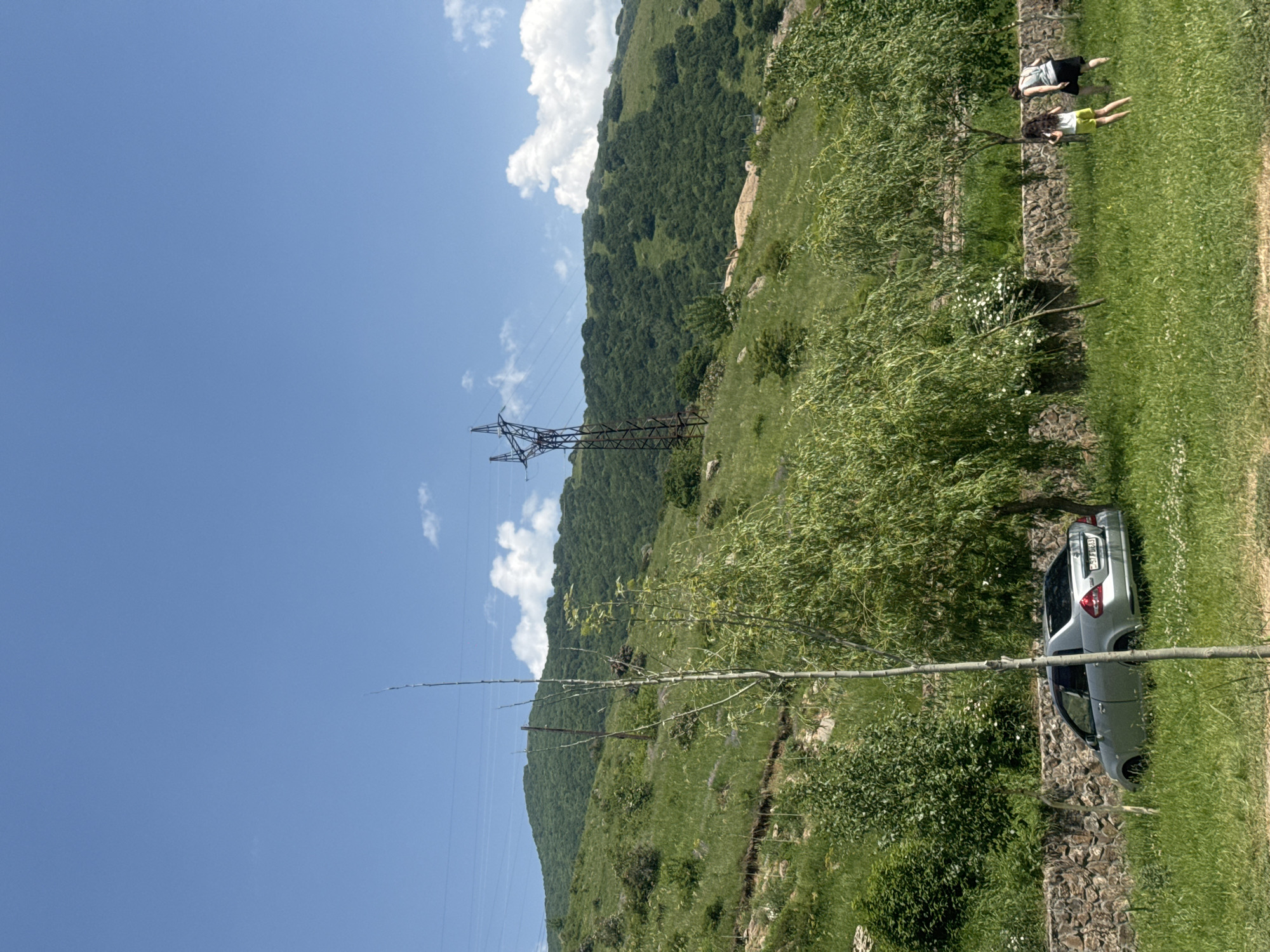 The image depicts a sunlit outdoor scene in Aghveran, Armenia. A silver sedan is parked on the left side of the frame, angled into the grassy area. In the mid-ground, two individuals, a woman and a girl, walk across the lawn away from the viewer. The background features a lush, green, tree-covered hillside under a clear blue sky dotted with white clouds. A tall, metal electricity pylon stands on the hillside, with power lines stretching across the frame. The foreground is characterized by a stone retaining wall bordering a grassy area with scattered trees and vegetation. The overall impression is one of a natural, rural landscape on a bright, clear day.