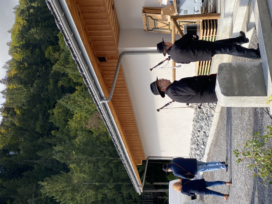 The image depicts an outdoor scene in Abondance, France, featuring a modern building against a backdrop of a dense, green forest covering a steep hillside under an overcast sky. The single-story building has a white facade, a dark tiled roof with light wooden eaves and soffit, and a grey metal gutter system. The ground in front of the building is covered with grey gravel, and a concrete barrier is positioned in the foreground.
Four individuals are present. On the right, two men are dressed in dark jackets, trousers, and dark hats. One man, with glasses and a beard, is observed in left profile. The second man is ascending concrete steps towards the building's entrance, his back largely to the viewer. Two bagpipes, featuring dark bags and light-colored chanters and drones, are propped vertically against the building wall near these men.
To the left of the frame, a woman with reddish-brown hair, wearing a dark jacket, blue jeans, and brown shoes, walks away from the camera. Adjacent to her, a man in a dark jacket, light blue jeans, and dark shoes also walks away, with a strap visible over his shoulder. A white commercial van is partially visible behind them on the far left. Stacked near the building's entrance are multiple wooden folding tables/benches, several cardboard boxes, and a white container.