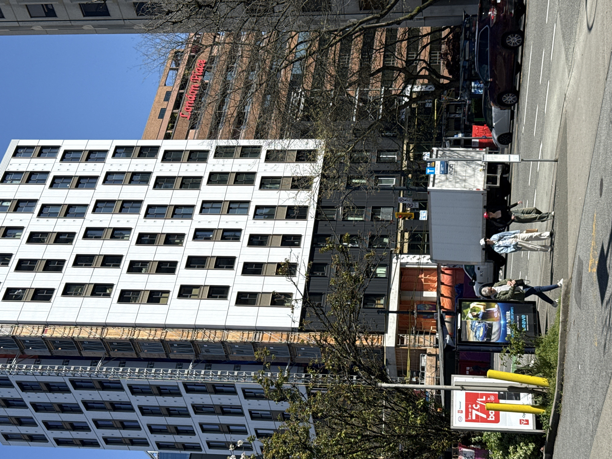 The image captures a vibrant street scene in Vancouver, Canada, under a clear, sunny sky, with a tilted perspective.
**Subjects:**
The foreground is dominated by a modern, multi-story building featuring a white and dark gray paneled facade with numerous rectangular windows. Scaffolding is visible along its lower section, indicating ongoing construction or maintenance. Behind it, a brown brick building features the red illuminated sign "London Place."
On the street level, several people are present. In the mid-ground, one person in a light striped shirt and light pants walks away from the camera, while another in a green jacket and glasses stands near a billboard. A third person with a red cap is seen further back near a white truck.
Vehicles include a dark SUV and a light-colored car parked along the curb, and a large white box truck and a smaller delivery truck on the road. Trees with bare branches or early leaves are interspersed throughout the urban landscape. Streetlights, traffic signals, and multiple billboards also line the street.
**Activity or event taking place:**
The scene depicts a typical bustling city day, with people walking along the sidewalk and various vehicles on the street, suggesting daily urban activities like commuting or errands.
**Notable details:**
The bright sunlight and clear blue sky indicate a daytime setting with pleasant weather. The mix of modern architecture and older buildings, along with visible construction, points to a dynamic urban environment. The sparse foliage on the trees suggests it might be early spring or late autumn.
**Visible text:**
* On the brown building: "London Place"
* On a billboard featuring a golfer: "BIGGER. BETTER. STREAM NOW. LIVE SPORTS. Crave." and "Rave."
* On another billboard: "Earn up to 7¢/L" and "Opti" (partially visible, likely part of a brand name), followed by "ba" (likely "back").
