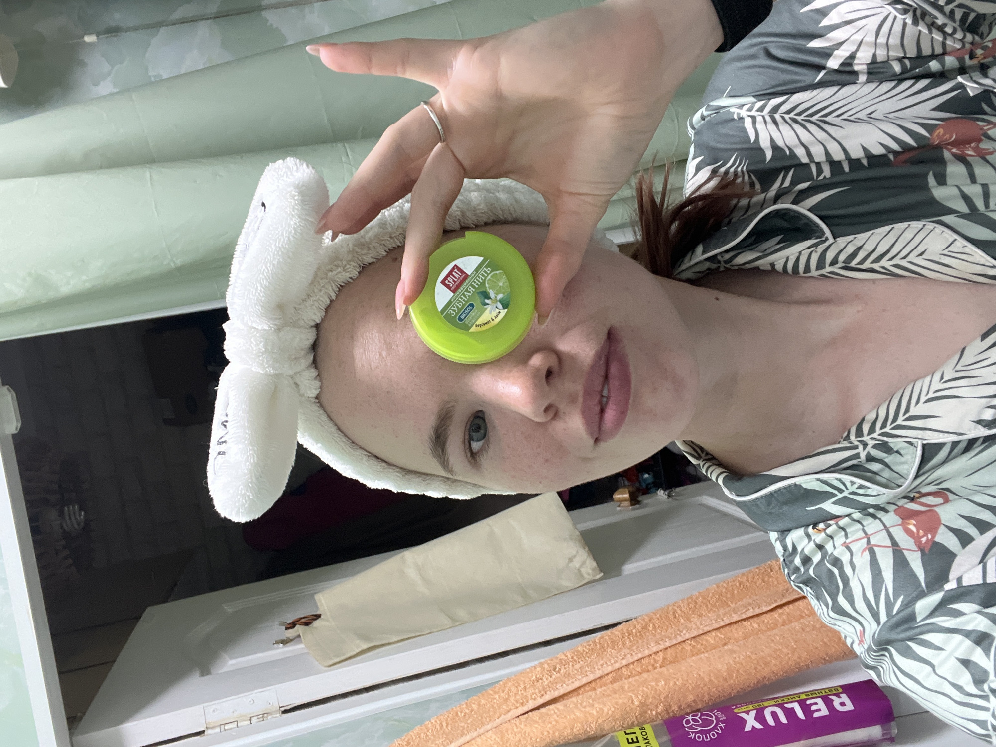 A young woman with light skin, freckles, and auburn hair tied back with a white fluffy headband is shown from the chest up, holding a small green container of dental floss to her right eye. She is wearing a pajama top with a tropical print of palm leaves and flamingos. The background appears to be a bathroom, with a white door and light green shower curtain visible. To the left of the woman, two folded towels are hanging on the door. The lighting is dim, and the overall impression is a casual, at-home selfie. The image does not provide any specific information about the city of Chaikovsky, Russia.