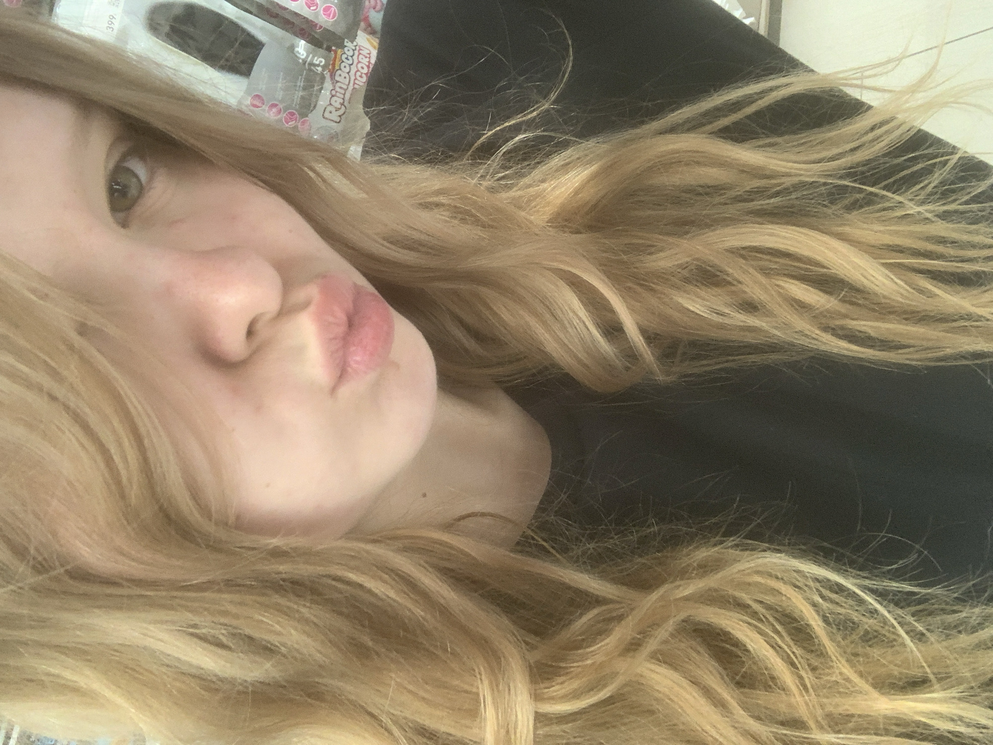 A close-up, selfie-style photograph depicts a young woman with long, wavy blonde hair. She has fair skin, light-colored eyes, and is pouting her lips. She is wearing a black top. In the background, partially obscured, are packaged goods, including a package labeled "Rainbocorns." The location is not discernible as being in Saint Petersburg, Russia.