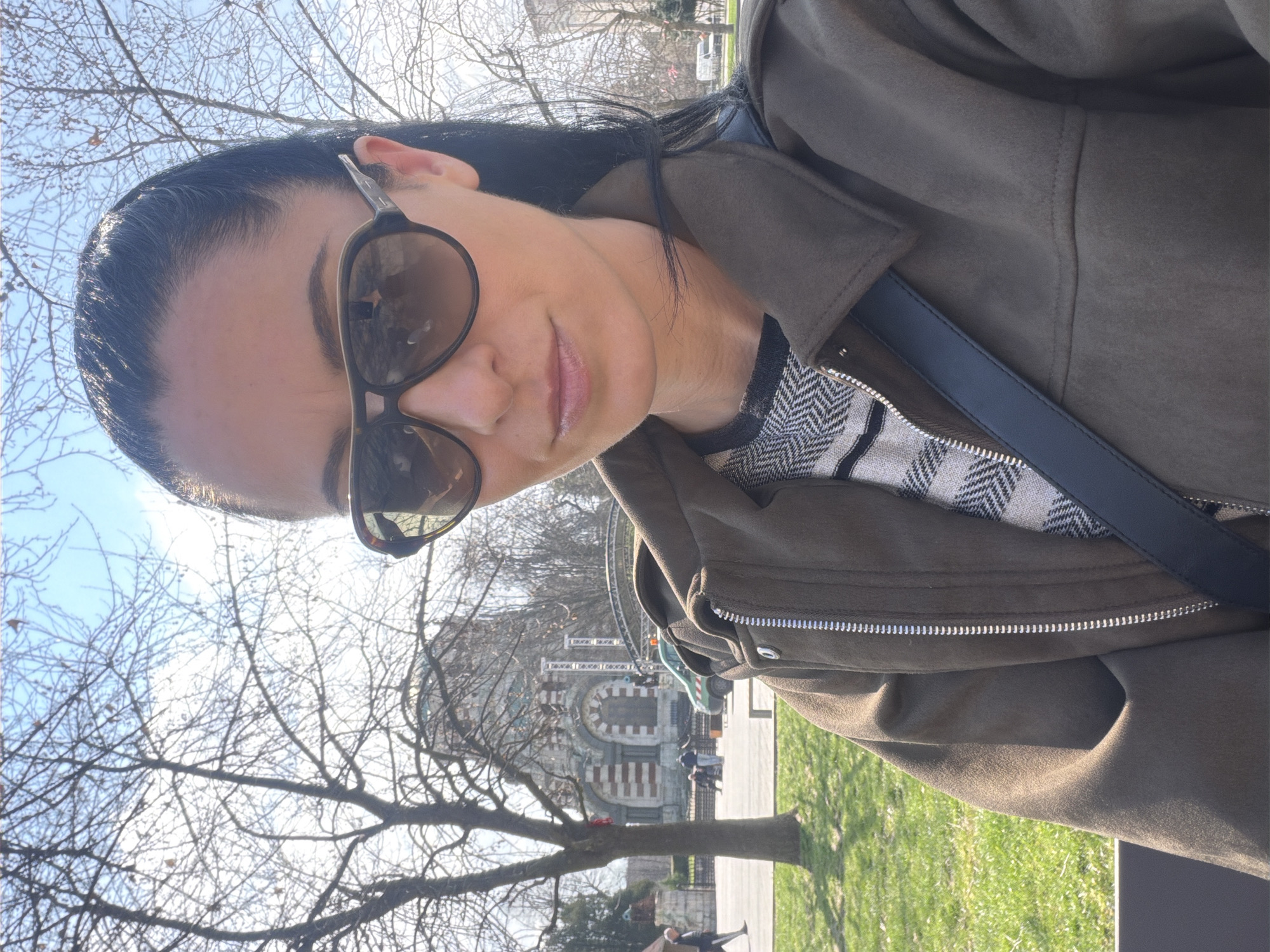 A woman with dark hair and sunglasses is the main subject of the image, wearing a dark jacket and a patterned top, facing the camera at an angle. The location appears to be a public park or area near the Alexander Nevsky Cathedral in Pleven, Bulgaria. In the background, there's a building that resembles a church or public building, typical architecture of Pleven. In the surrounding area, there are trees, grass, and other individuals visible in the background. The trees appear to be bare, indicating that it's either early spring or late fall.