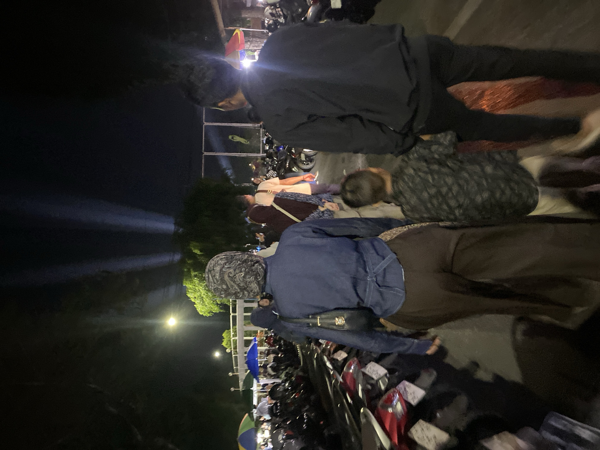 The image depicts a nighttime scene in what appears to be a parking area or a street in Banjarbaru, Indonesia. In the foreground, a group of people are walking away from the camera. The central figure is a person wearing a dark blue denim-like top and dark brown pants, with a headscarf. To their right, a child in a patterned shirt and light-colored pants walks alongside. Further to the right, a person in a black jacket and black pants is also walking away. In the background, numerous motorcycles are parked, suggesting this is a public area where people have arrived by vehicle. Several colorful umbrellas are visible among the parked motorcycles, possibly indicating stalls or temporary shelters. Overhead, spotlights cast beams of light into the dark sky, and trees with green foliage are present. The overall atmosphere suggests a lively evening event or gathering.