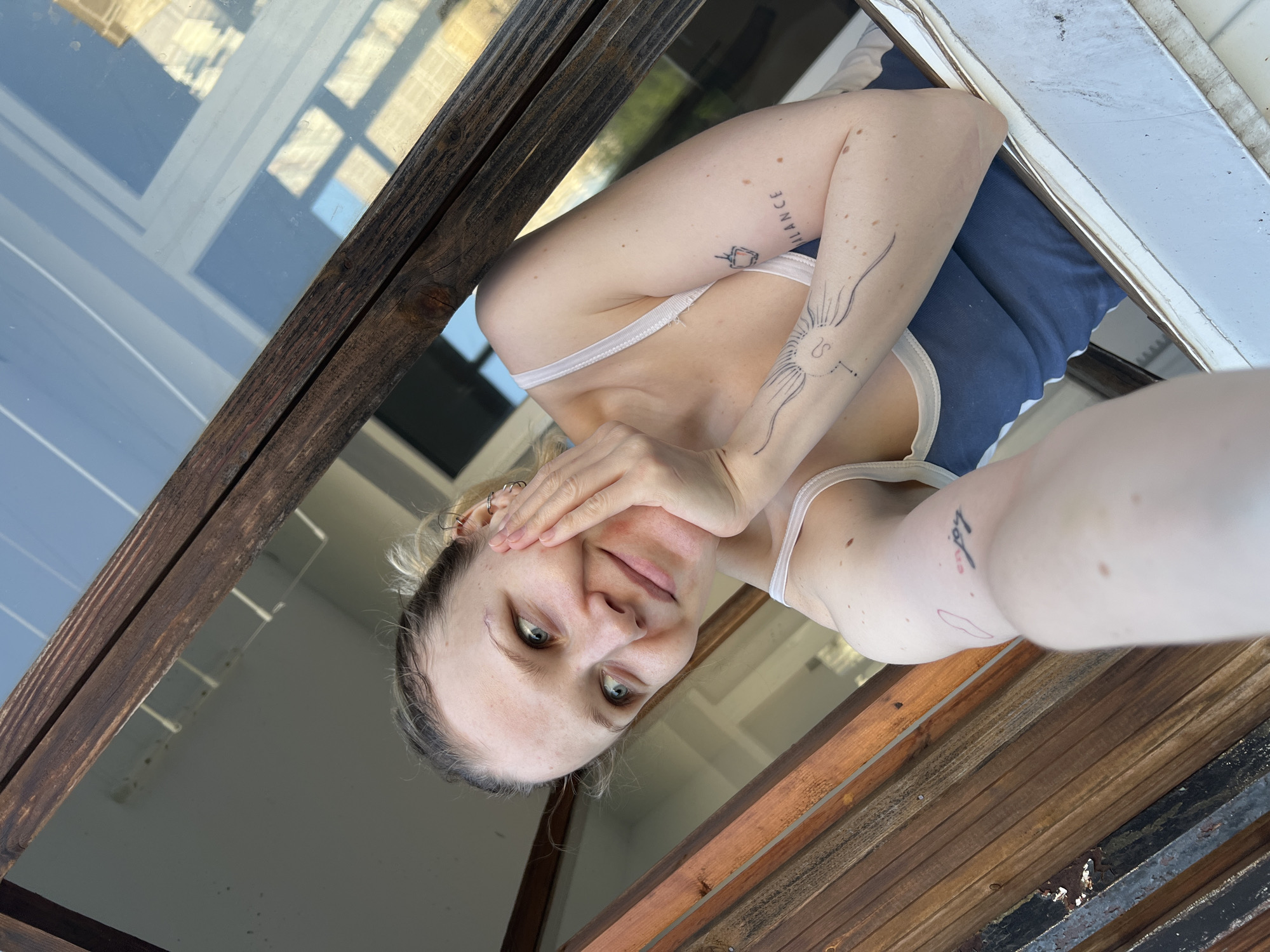 A selfie of a woman taken from a low angle, positioned within a wooden-framed window. She is fair-skinned with blonde hair pulled back, and has tattoos on her arms. She is wearing a white tank top. The window appears to be in Khimki, Russia based on metadata.