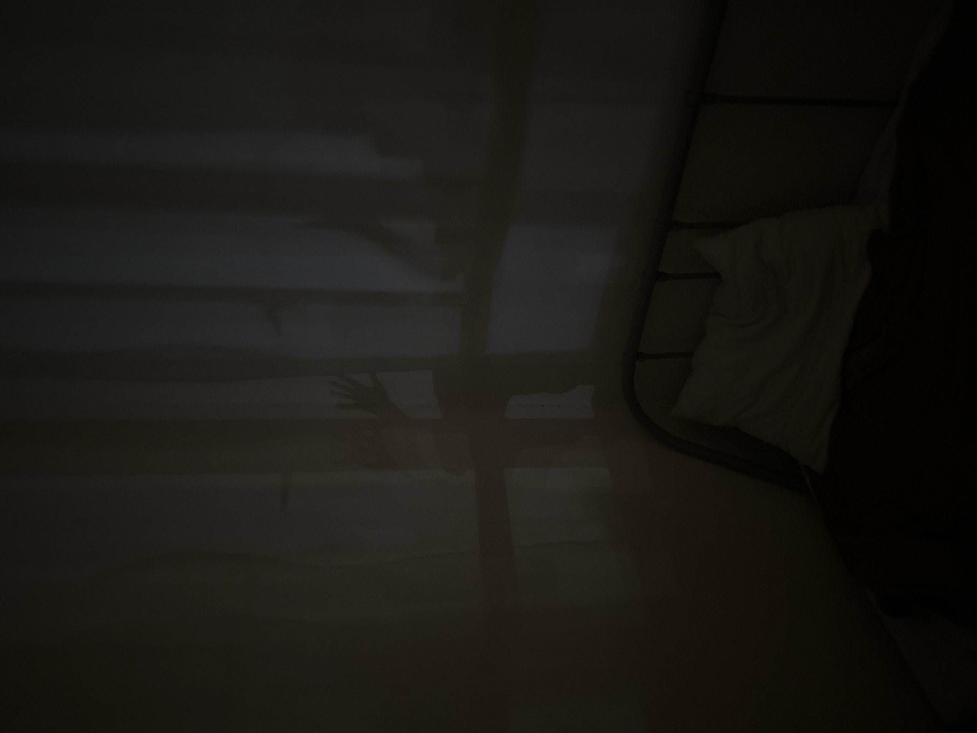 The image depicts a dimly lit bedroom with a single bed. Shadows cast by a window with sheer curtains create an eerie atmosphere. The shadows suggest the presence of a person or persons. A pillow rests on the bed, and a dark blanket covers the lower portion. The bed frame is visible. The overall impression is one of unease and mystery. The location in Sinyavino, Russia, is not explicitly depicted in the image itself but is provided as context.