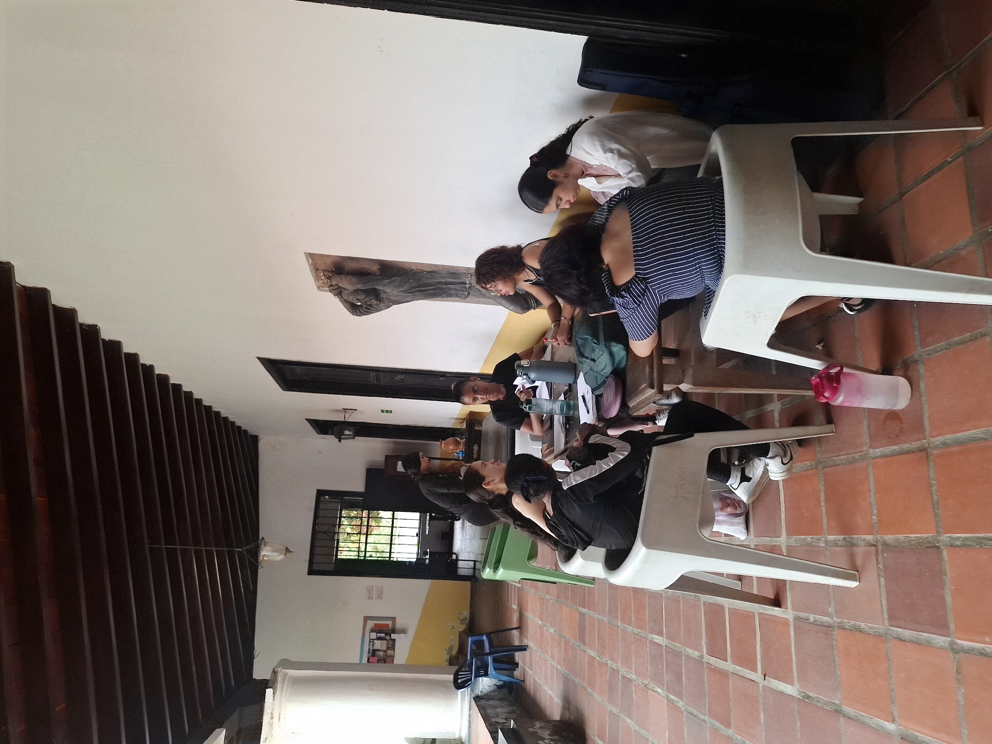 The image shows a group of people seated indoors. They appear to be focused on a table or some activity in front of them. The location may be in El Valle del Espíritu Santo, Venezuela, judging from the prompt, but the image does not provide visual confirmation. The floor is covered with red tiles and there are chairs made of plastic. There is a dark wooden ceiling with slats on the left side of the image, and a window.