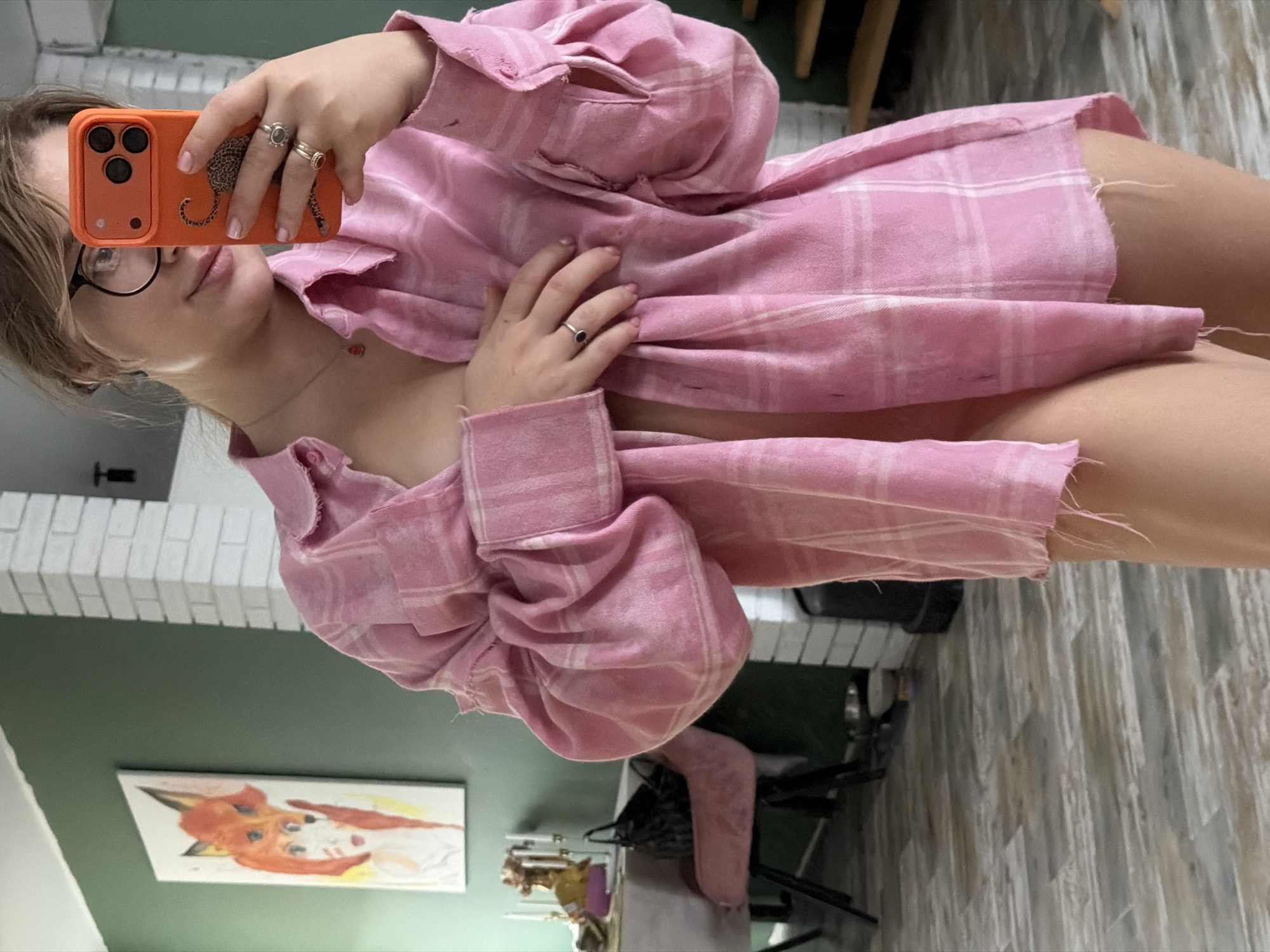 An indoor mirror selfie features a young individual, likely female, taking a picture with an orange smartphone. The person has light brown hair, wears black-rimmed glasses, and has light pink nail polish on their fingers. Several rings adorn their fingers, including some with dark stones. A delicate necklace with a small pendant is visible around their neck.
The person is wearing a loose-fitting, oversized pink plaid shirt or jacket, which appears somewhat distressed or raw-edged at the seams, suggesting a casual or worn style. Their right hand is resting on their chest, slightly pulling the fabric of the shirt. The orange phone case features a distinct black leopard print design.
The background of the image reveals an indoor setting, likely a room in a home. A green wall is visible behind the person, topped by a white brick-patterned structure, possibly a fireplace mantle or a decorative architectural element. On the green wall, a framed picture depicts a stylized, reddish-brown and white fox or dog character. Another abstract painting is partially visible further in the background. The flooring appears to be a light-colored, possibly patterned rug or wood. Furniture elements like a chair with a pink fabric component are also present. The overall scene suggests a personal and relaxed environment, captured in Большая Шильна, Russia. No discernible text is visible in the image.