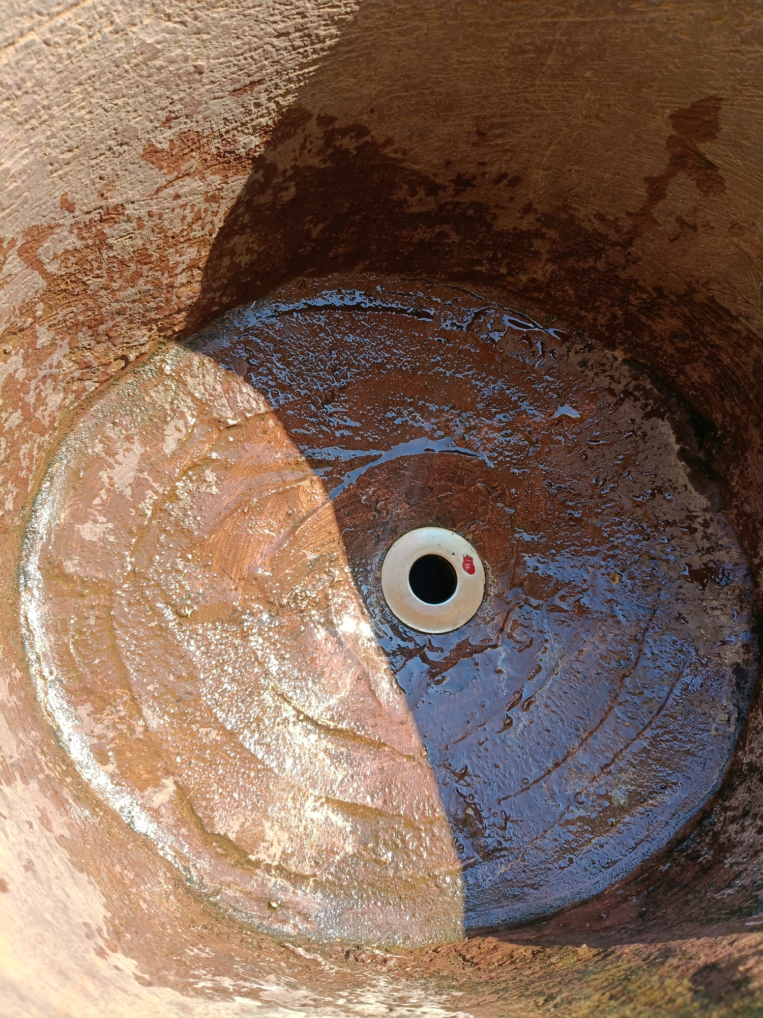 This is a top-down, close-up shot of the inside of a large, reddish-brown clay pot. The pot appears weathered, with textured, uneven surfaces. A portion of the pot is dry and lighter in color, while the other half is wet and significantly darker. Water is pooled in the wet section, forming small, shimmering droplets and reflecting light. In the center of the wet area is a circular, metallic drain with a black opening. The transition between the dry and wet areas is sharp, suggesting a recent exposure to water or a deliberate washing. A diagonal shadow cuts across the pot, indicating the presence of sunlight from the right. The overall impression is one of a functional, possibly rustic, utilitarian object.