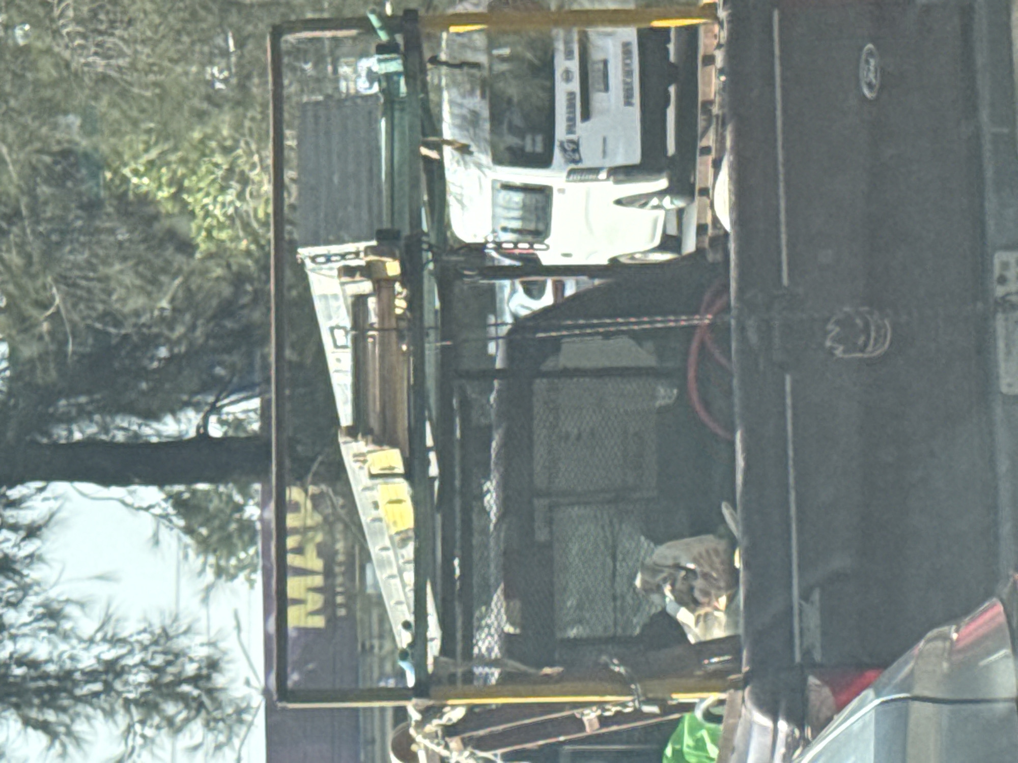 The image depicts the back of a truck filled with various items, possibly waste or construction material. A white Subaru Forester bearing the word "PROTECCIÓN" is parked in the background. A ladder rests against the side of the truck bed. The environment suggests an urban or suburban setting with trees and commercial signage visible. The sign reads "MAK... UNIVERSIDAD". Given the context clues of the word "UNIVERSIDAD" (University), the location is in the city of Puebla, Mexico.