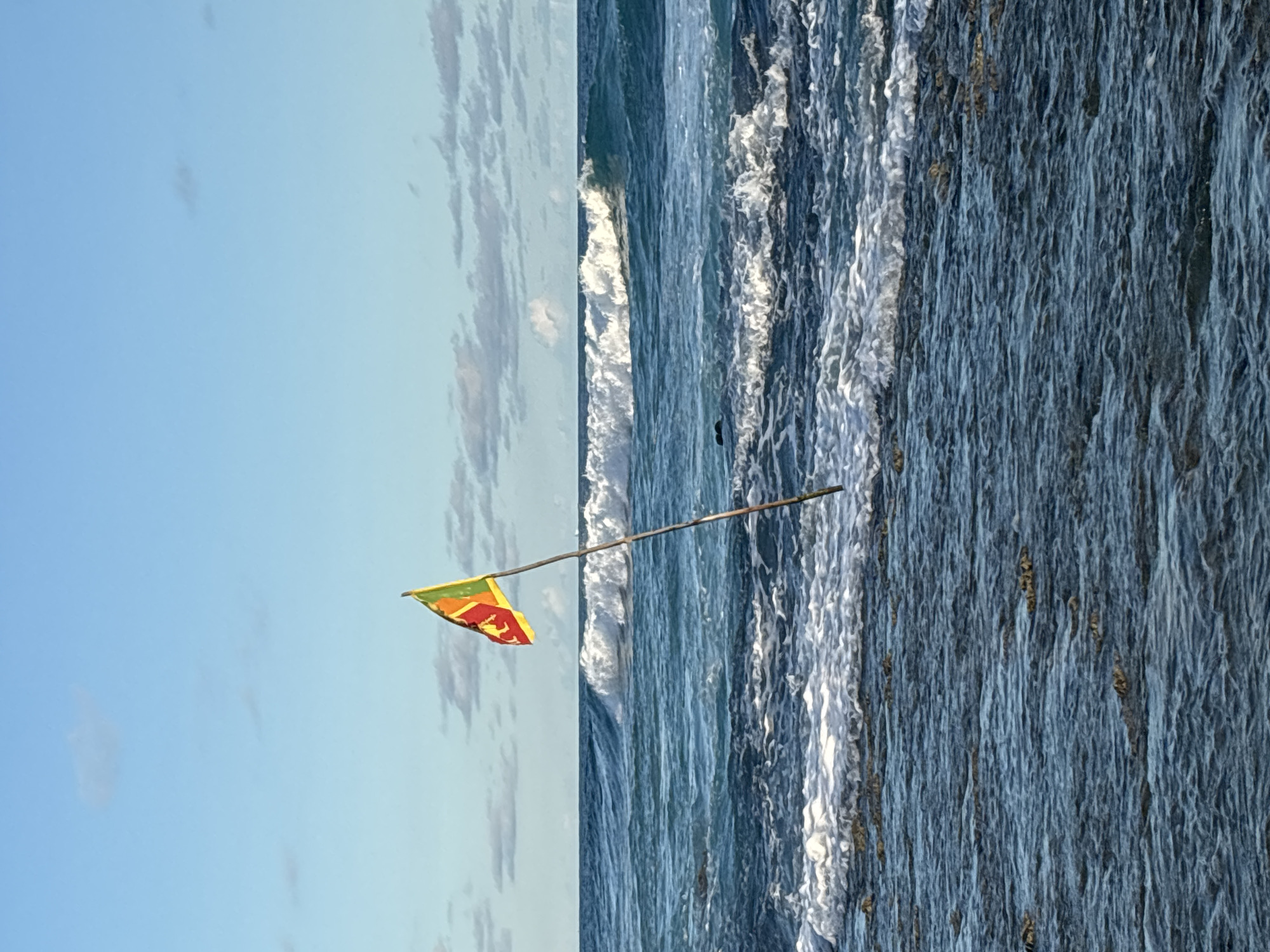 A low-angle shot of the Sri Lankan flag attached to a stick, with a blue sky containing thin clouds on the left side, and the ocean on the right side. The ocean water contains visible foam. The photo was taken in Thalaramba, Sri Lanka.