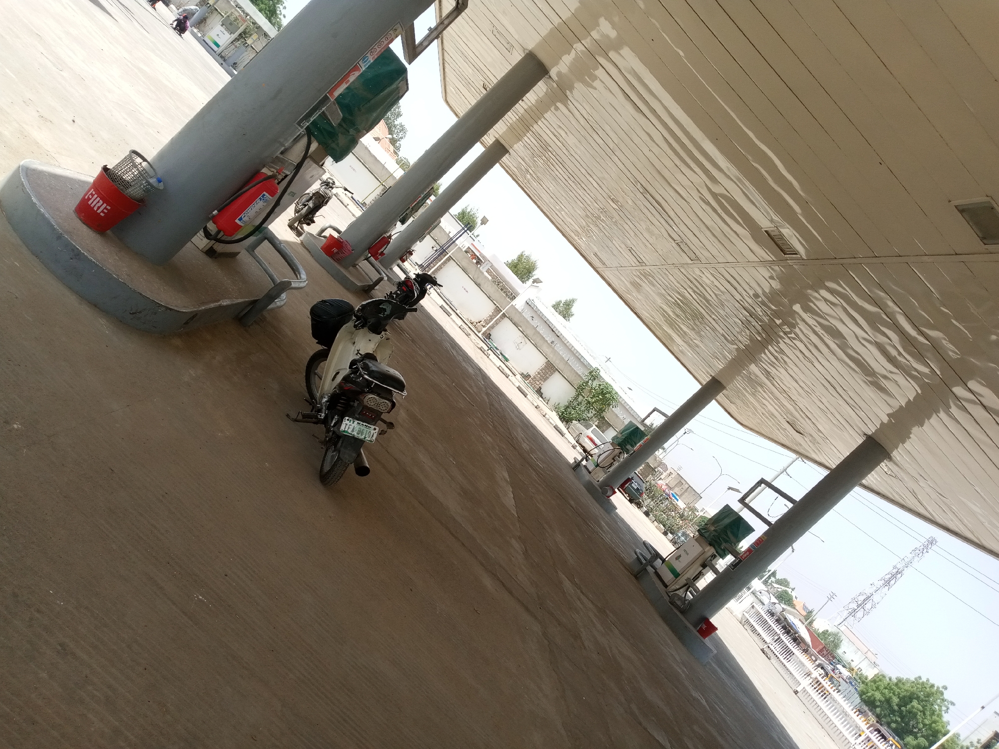 The image depicts a gas station scene with a motorcycle parked under a covered canopy. Several gas pumps are visible, some with green covers. A red fire bucket with "FIRE" written on it in white letters is positioned near one of the support poles. A red fire extinguisher is also mounted on a pole. The ground is paved with concrete. In the background, there are buildings and trees, suggesting an outdoor, possibly urban or suburban, setting. A tall communication tower is visible in the distance. The lighting suggests it is daytime, with bright sunlight. The overall impression is a functional, everyday scene at a gas station. The motorcycle has a license plate with some visible text.