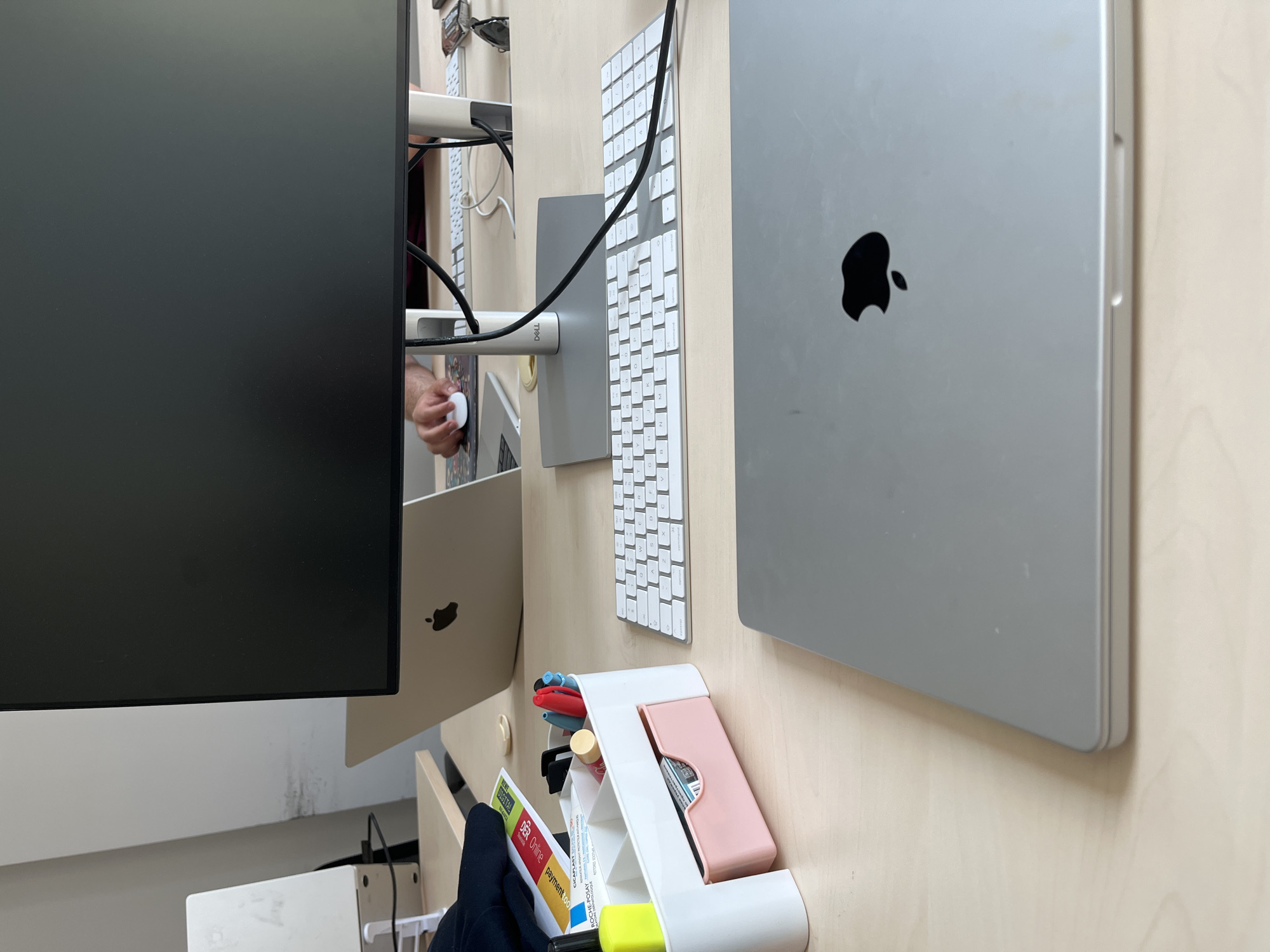 The image shows a desk with a person's hands using a computer. The subjects are primarily computer equipment, including a large black monitor on the left, a silver Apple laptop in the center right, and a white Apple keyboard positioned between them. There are also some desk accessories in the foreground, such as a white organizer with pens and markers, and a small pink case. A person's hands are visible, one holding a white computer mouse, and the other reaching towards the keyboard. The scene appears to be an indoor workspace. The lighting suggests it could be daytime, but there are no clear external cues. The overall impression is of someone working or browsing on a computer. The presence of Apple products suggests a modern, tech-oriented environment. There is some visible text on a document in the organizer, including "DER Online" and "payment.co".