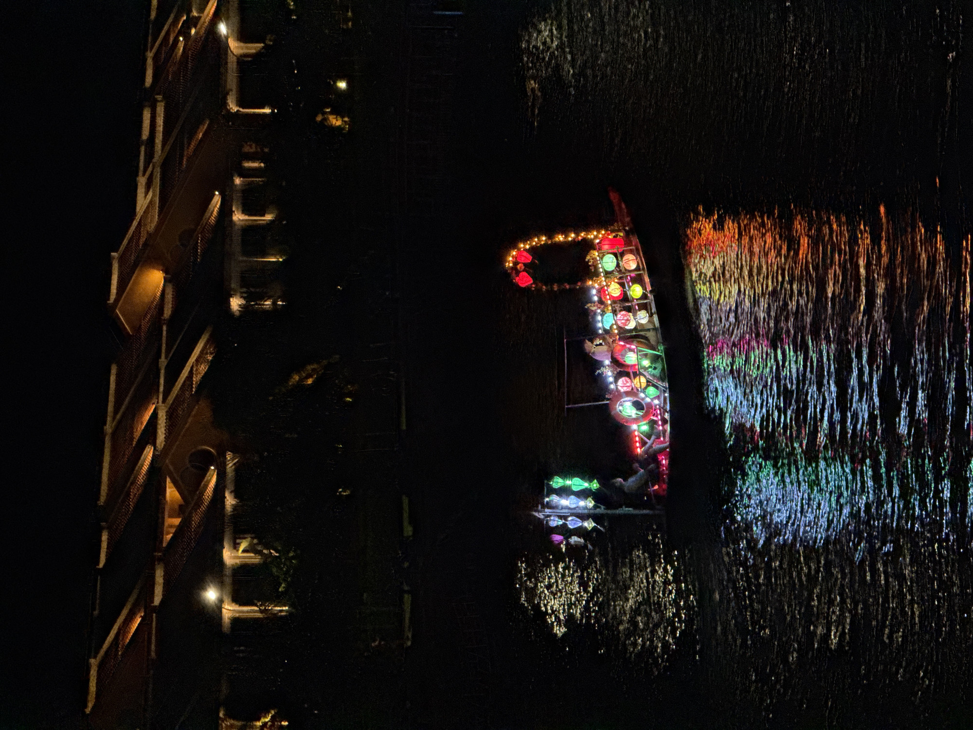 In Hoi An, Vietnam, at night, a boat adorned with colorful lanterns navigates a dark river. The lanterns, in shades of green, pink, yellow, and red, are strung across the boat, illuminating its interior and casting reflections on the rippling water. A figure is seated on the boat. In the background, a row of houses with tiled roofs is visible, their windows and eaves softly lit by warm interior lights, suggesting a residential area. The scene captures a tranquil and festive atmosphere.