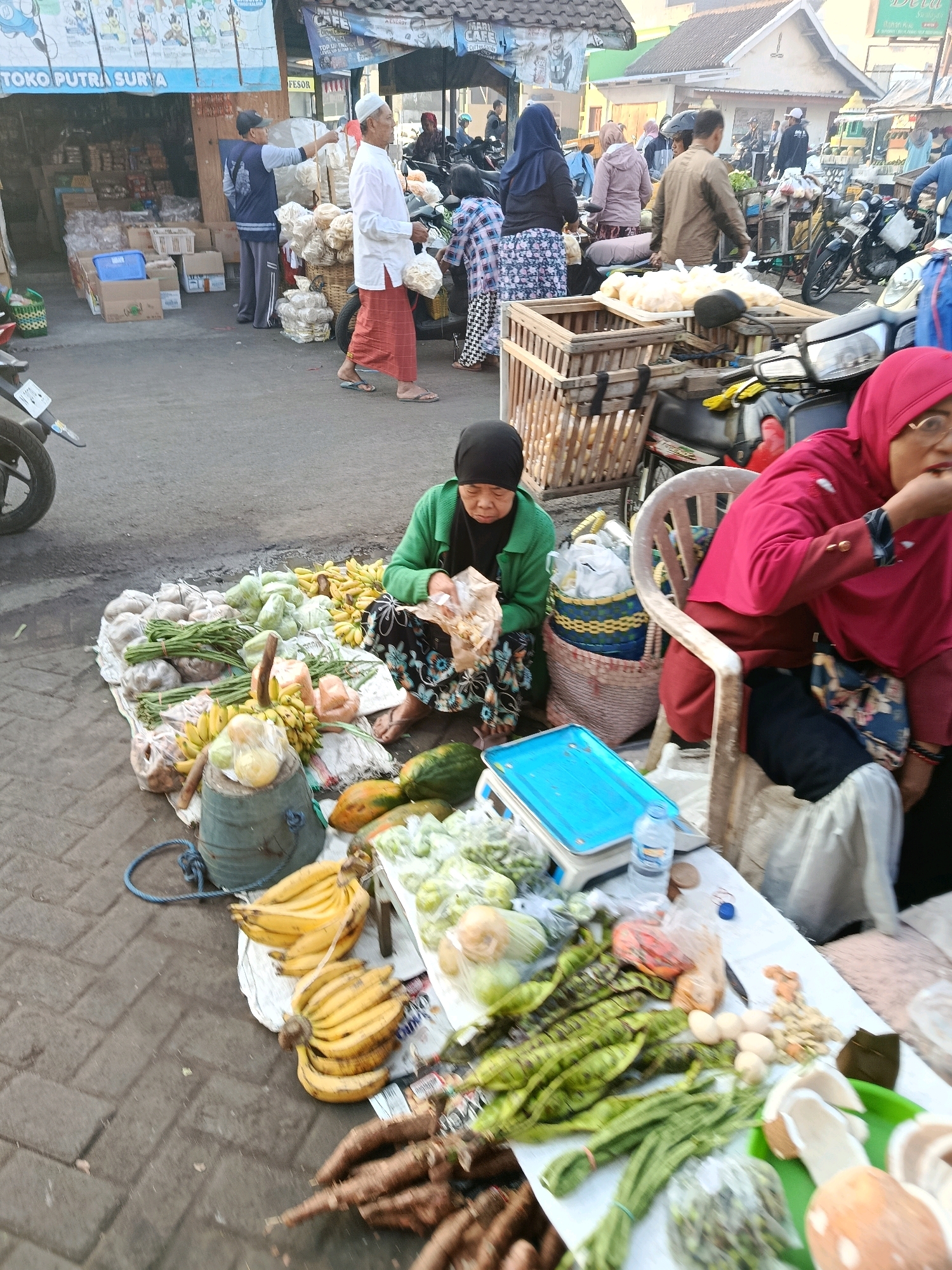 A bustling street market scene unfolds in Bululawang, Indonesia, with various individuals engaged in commerce. The foreground features a woman in a green cardigan and a floral skirt, seated on the ground and handling food items, likely for sale. Next to her, a woman in a vibrant red hijab and matching clothing sits on a plastic chair, also appearing to be attending to customers or food. A variety of fresh produce, including bananas, leafy green vegetables, long beans, cassava, and what appear to be coconuts, are displayed on a white tarp and a green tray, indicating a food vendor's stall.
Behind them, a wooden cart filled with goods, possibly more produce or merchandise, sits beside a motorcycle. Several other individuals are visible in the background, including a man in a white shirt and sarong, holding a bag of what looks like crackers, and another man in a dark blue shirt and a cap, gesturing towards a stall. A stall with a blue awning labeled "TOKO PUTRA SURYA" is seen to the left, suggesting a general store. The ground is paved with brick-like stones, and the overall atmosphere suggests a busy morning or afternoon at a local Indonesian market. Motorcycles and other market stalls are present, contributing to the lively and crowded environment.