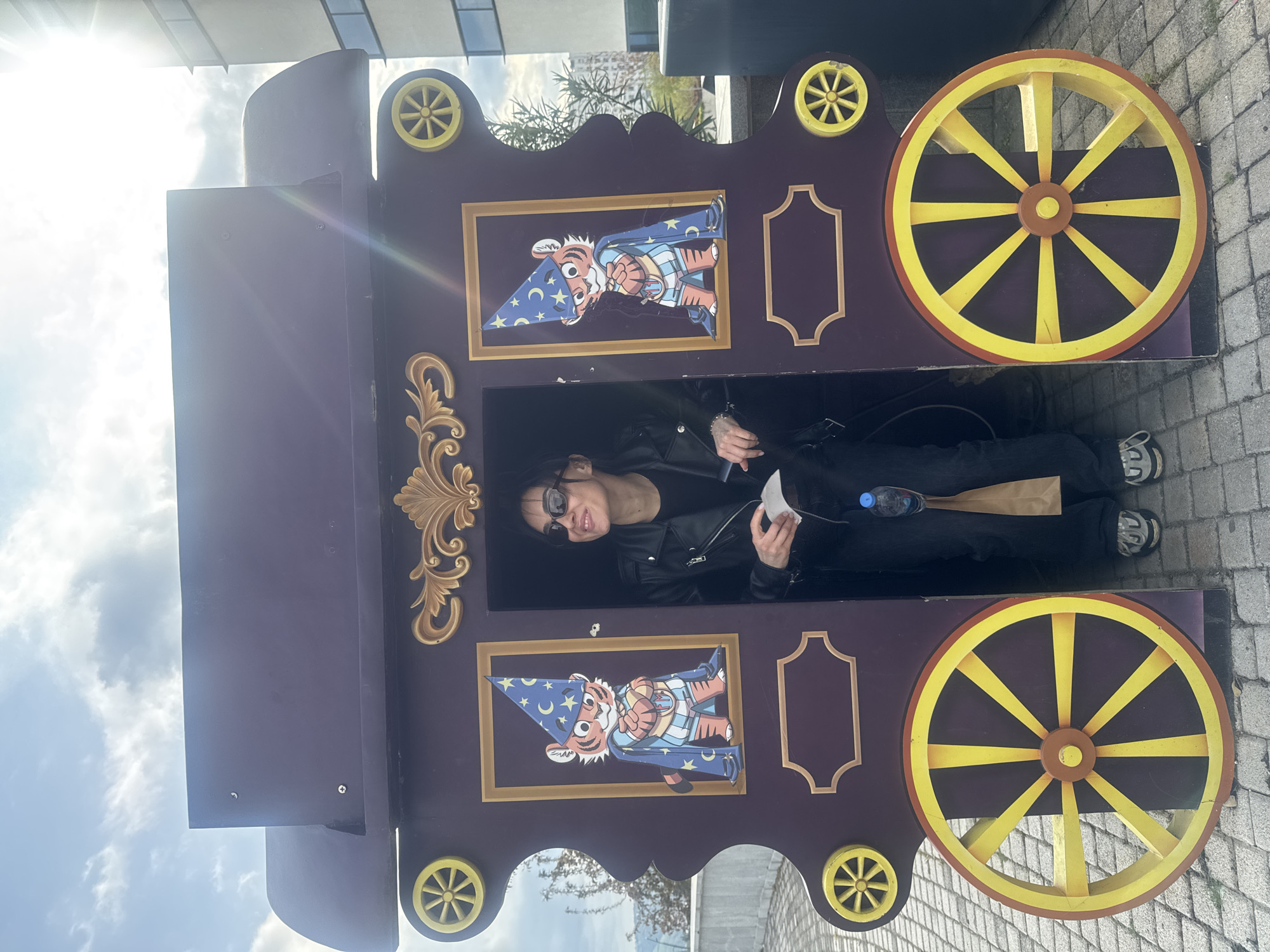 A woman is posing within a decorative, purple wooden wagon or cart-shaped photo stand outdoors on a bright day. The wagon features prominent yellow wooden wheels, smaller yellow decorative wheels on its upper frame, and ornate gold scrollwork. Within two framed sections on the wagon, there are cut-outs of a cartoon tiger dressed as a wizard, complete with a blue hat adorned with stars and a matching blue cape.
The woman is lying down inside the wagon's opening, looking towards the camera with a smile. She is wearing a black leather jacket, dark trousers, sunglasses, and white and grey sneakers. She appears to be holding a piece of white paper or tissue, and a blue plastic water bottle and a brown paper bag are visible near her.
The setting includes a paved ground, possibly cobblestones or bricks. A bright sky with some clouds is visible, and a strong sun flare is prominent in the upper left of the image, indicating sunny weather. Portions of buildings are faintly visible in the background. The overall impression is a cheerful, casual scene. No discernible text is visible in the image. The location is contextualized as Tbilisi, Georgia.
