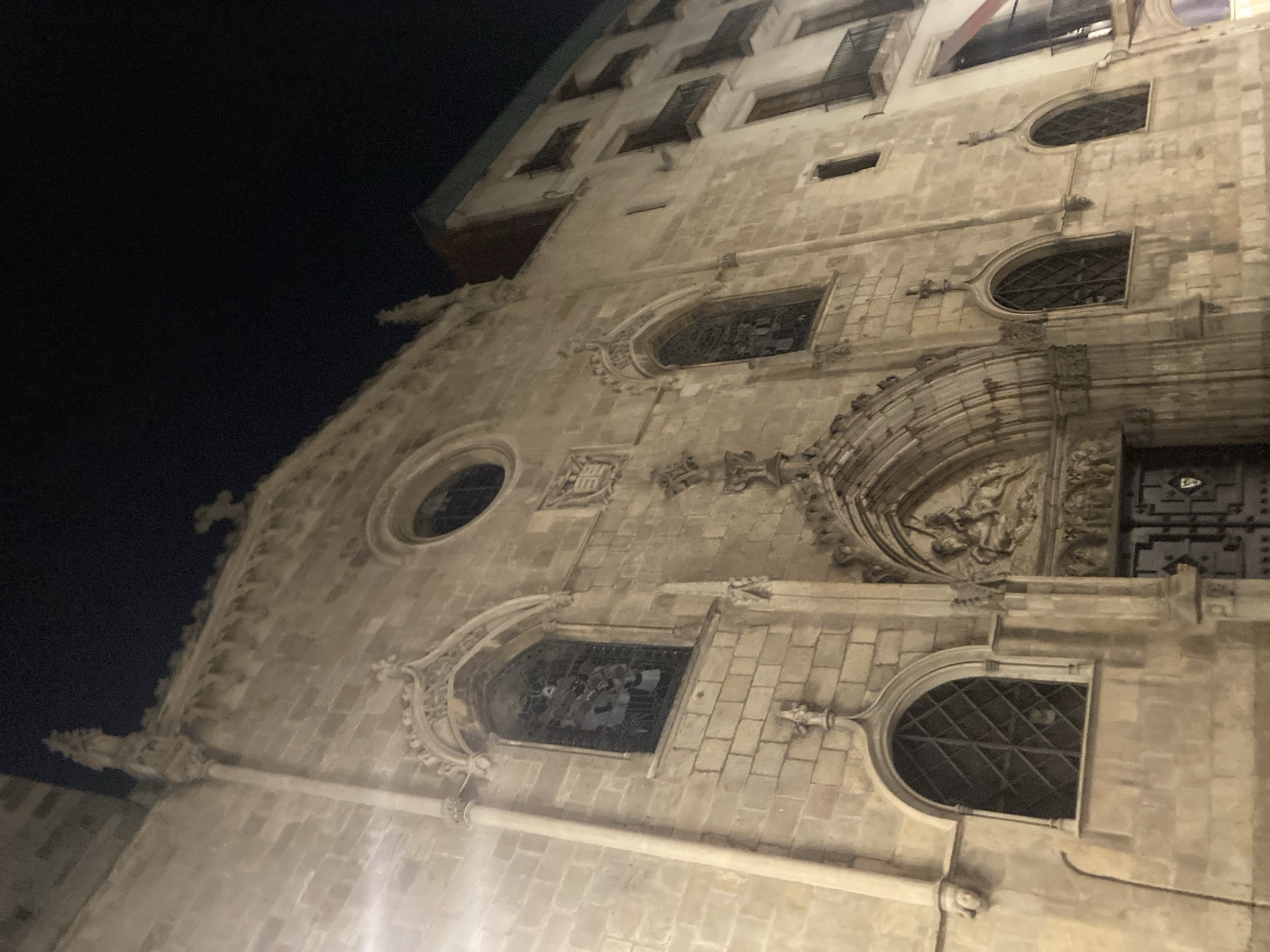 The image shows the facade of a stone building, likely a church, at night in Barcelona, Spain. The building features Gothic architectural elements such as pointed arches, ornate carvings above the entrance, and stained glass windows. Several windows are visible, some with intricate metal grilles. Above the entrance, a relief sculpture depicts a scene with figures, including a horseman. To the right of the church, a modern apartment building with balconies and windows is visible. The sky is dark, indicating it is nighttime. The lighting suggests artificial illumination, possibly from streetlights or the building itself, casting a warm glow on the stone. No people are clearly visible in the frame.