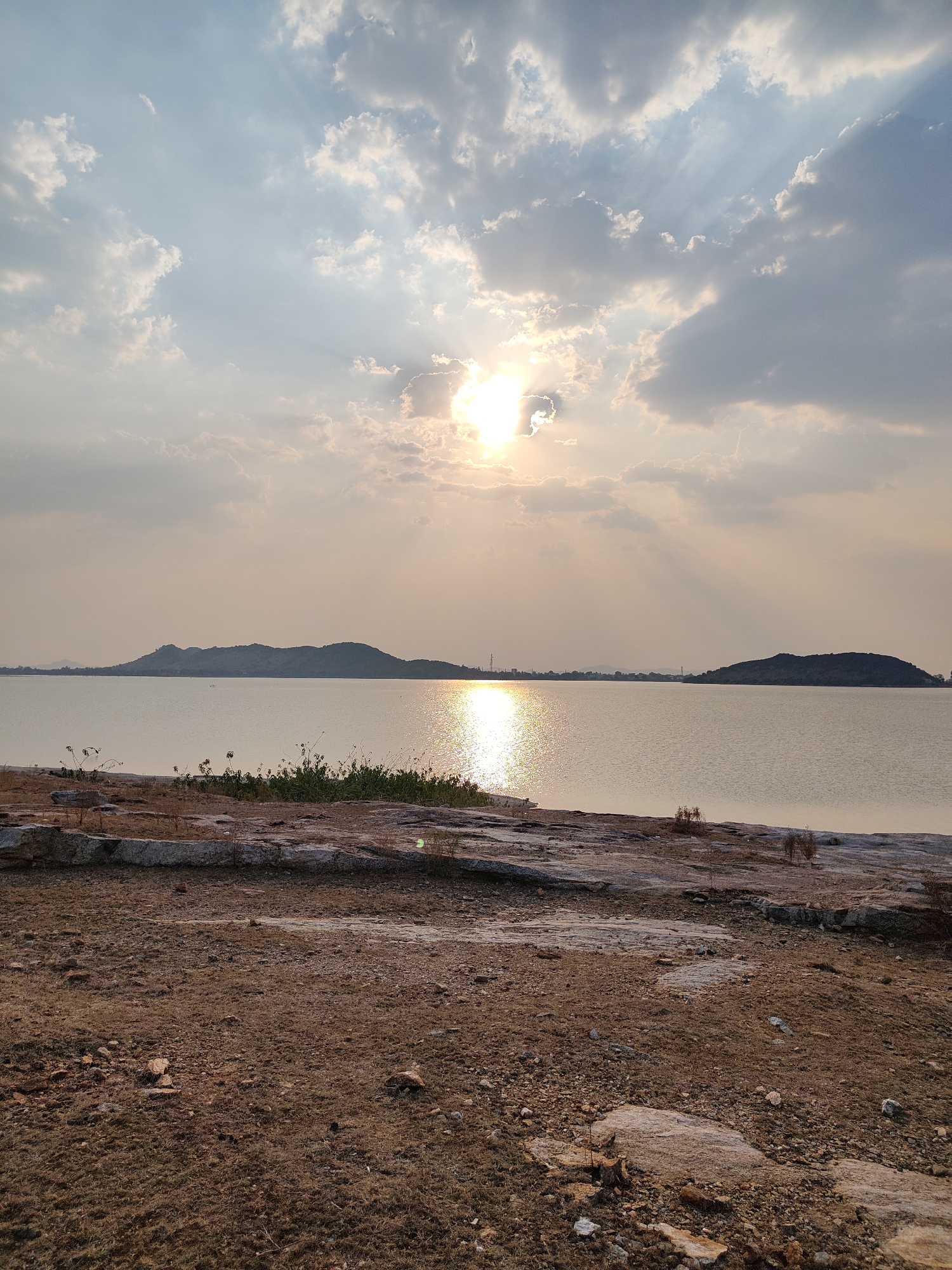 The image depicts a tranquil scene at a lake in Kammavaripalli, India, under a sky filled with clouds partially obscuring the sun. The sun's rays stream down, creating a shimmering reflection on the water's surface. Two landmasses appear in the distance. The foreground features dry, rocky terrain leading to the water's edge. Sparse vegetation is visible near the waterline. There are no people visible.