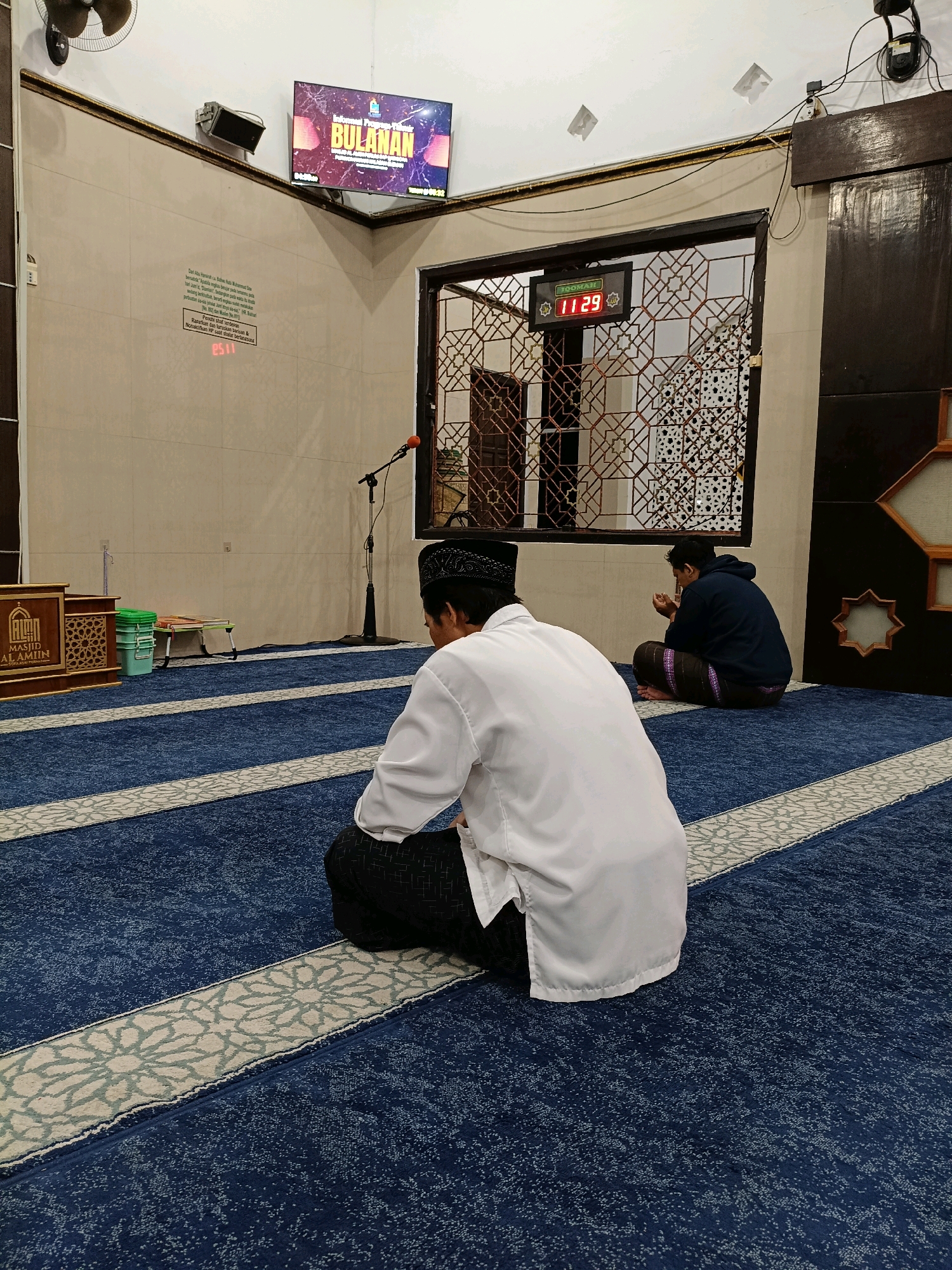 The image depicts the interior of a mosque in Maguwoharjo, Indonesia, with a blue carpet covering the floor, adorned with white and gray patterned stripes. Two men are visible, both seated on the carpet facing away from the camera. The man in the foreground wears a white long-sleeved shirt and black pants, with a black peci cap. The man in the background wears a dark blue hoodie and a patterned sarong.
In the background, a digital clock displays "11:29" and "JOOMAH". A large screen mounted on the wall shows an image with the text "Informasi Program Tahunan BULANAN". A microphone on a stand is positioned in the middle ground. To the left, a wooden podium with an inscription is visible. Various objects, including a green bucket and a small foldable table, are also present. The walls are tiled, and a decorative screen with geometric patterns is visible. A ceiling fan is mounted in the upper left corner of the image.