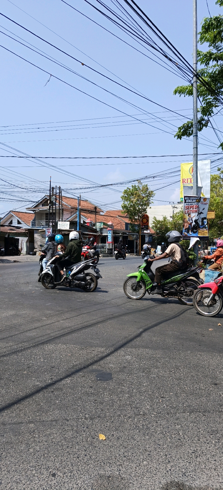 The image depicts a street scene in Kebumen, Indonesia, likely at an intersection given the presence of multiple vehicles and the road markings. The sky is clear and blue, with a complex network of overhead power lines crisscrossing the frame. In the foreground, the asphalt road is visible, with shadows cast by the power lines.
Several motorcycles are in motion. On the left, a white scooter carries three people, two of whom are wearing helmets. One rider wears a dark jacket and jeans, and the passenger behind them wears a light-colored shirt. A third person is partially obscured behind them. Further to the right, a person on a green motorcycle with black accents is visible, wearing a light brown shirt, dark pants, and a grey helmet, with a backpack on their back. A red motorcycle with a pink-seated rider is positioned to the right of the green motorcycle, partially cropped from view. In the background, other motorcycles are seen in motion or parked.
Buildings line the street, characterized by orange tiled roofs and light-colored walls. Some buildings appear to be shops or commercial establishments, with signs visible, including a yellow banner with Indonesian text and a poster advertising electric motorcycles. Trees with green foliage are present, particularly on the right side of the image, and a utility pole with multiple cables is also visible. The overall impression is of a busy, everyday street scene in a tropical urban environment.