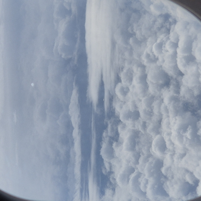 The image displays an aerial view captured from an aircraft window, indicated by the curved dark borders on the left and right edges. The scene is dominated by a vast, textured expanse of white cumulus clouds, forming an undulating layer across the lower and mid-sections of the frame. Above this dense cloud cover, lighter, more scattered cloud formations and sections of a pale blue sky are visible.
Two distinct objects are present within the aerial environment. In the upper-central portion of the image, a bright, reflective, somewhat circular or oval-shaped object is visible against a lighter cloud background. Below this, nearer the horizontal center of the frame, a smaller, dark, elongated object, appearing as a horizontal line with minimal structural detail, is observed within a thinner cloud layer. This observation is made from an aircraft in the vicinity of Blagnac, France.