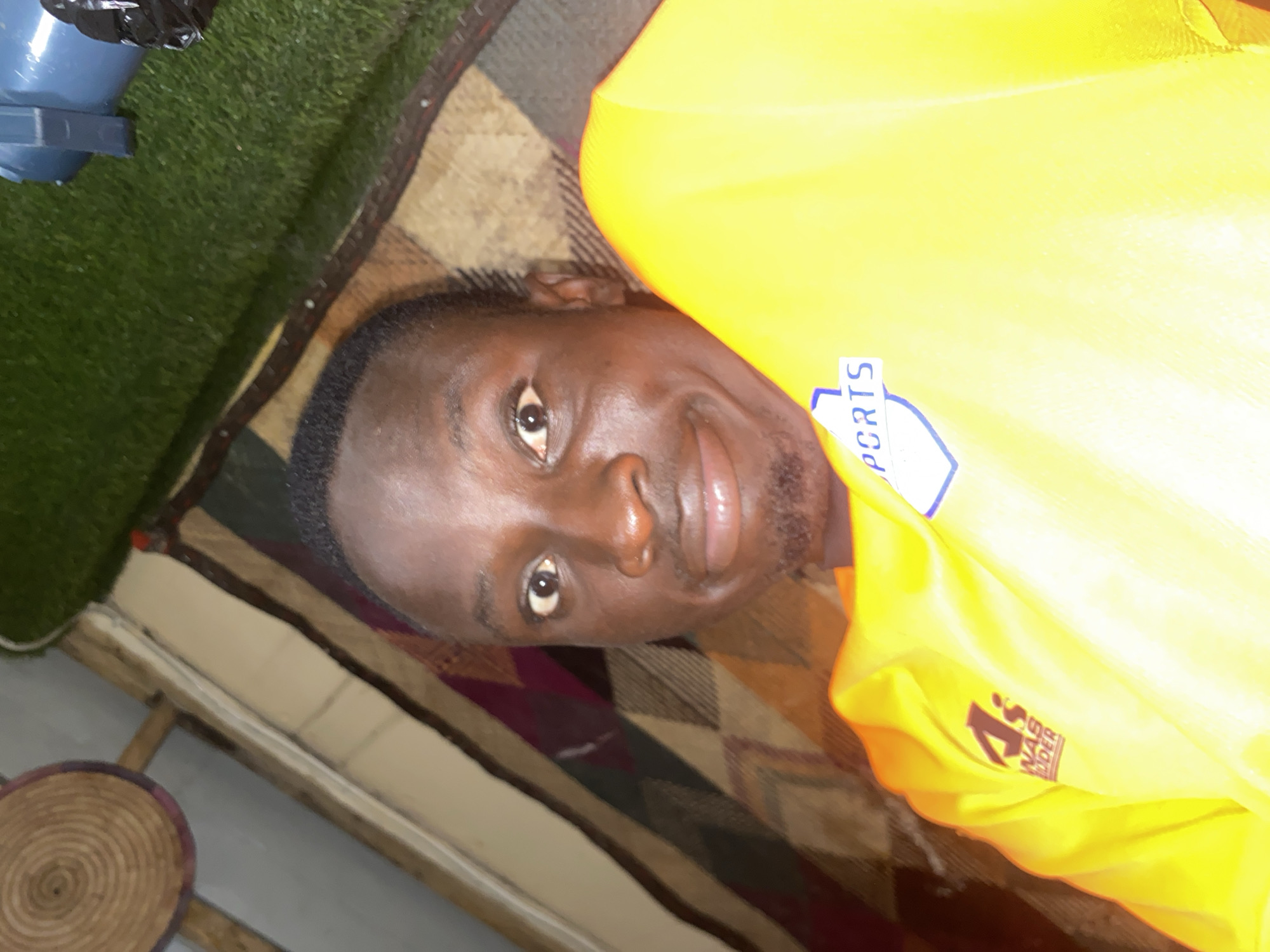 The image is a selfie taken by a dark-skinned male lying down. He is wearing a bright yellow shirt with a blue and white shield logo on the left chest that reads "SPORTS" and another logo on the left sleeve that appears to be a stylized "A" with text below it. The background is a mix of patterned fabric, possibly a blanket or rug, and a patch of artificial green turf. A woven circular object is visible in the upper left corner, and a blue object, possibly a light fixture or part of a machine, is in the upper right corner. The lighting suggests an indoor setting, possibly at night or in a dimly lit area. There is no visible indication of the specific location within Barden Yana, Nigeria.
